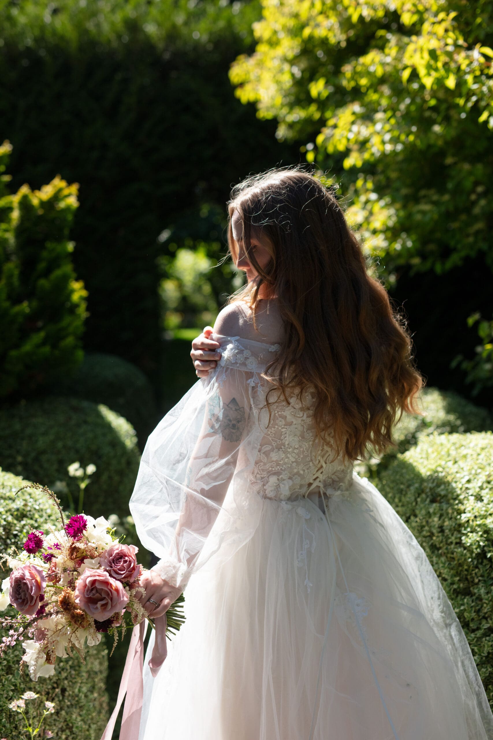 Bride in a flowing gown holds a textured bouquet in a sunlit garden, caught in a quiet, reflective moment before the ceremony.