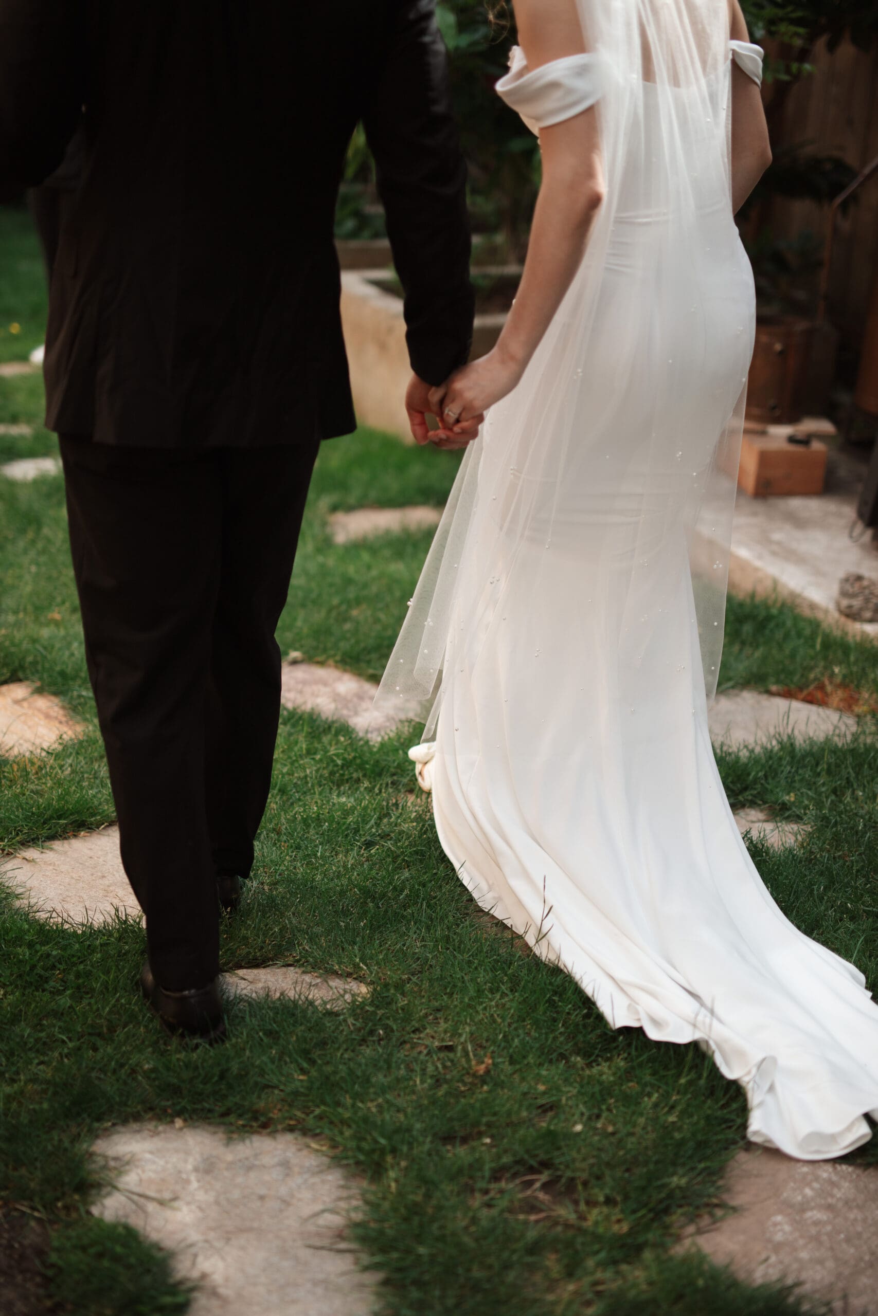 Close-up of bride and groom walking hand-in-hand across a stone path, her fitted dress and veil catching soft afternoon light.