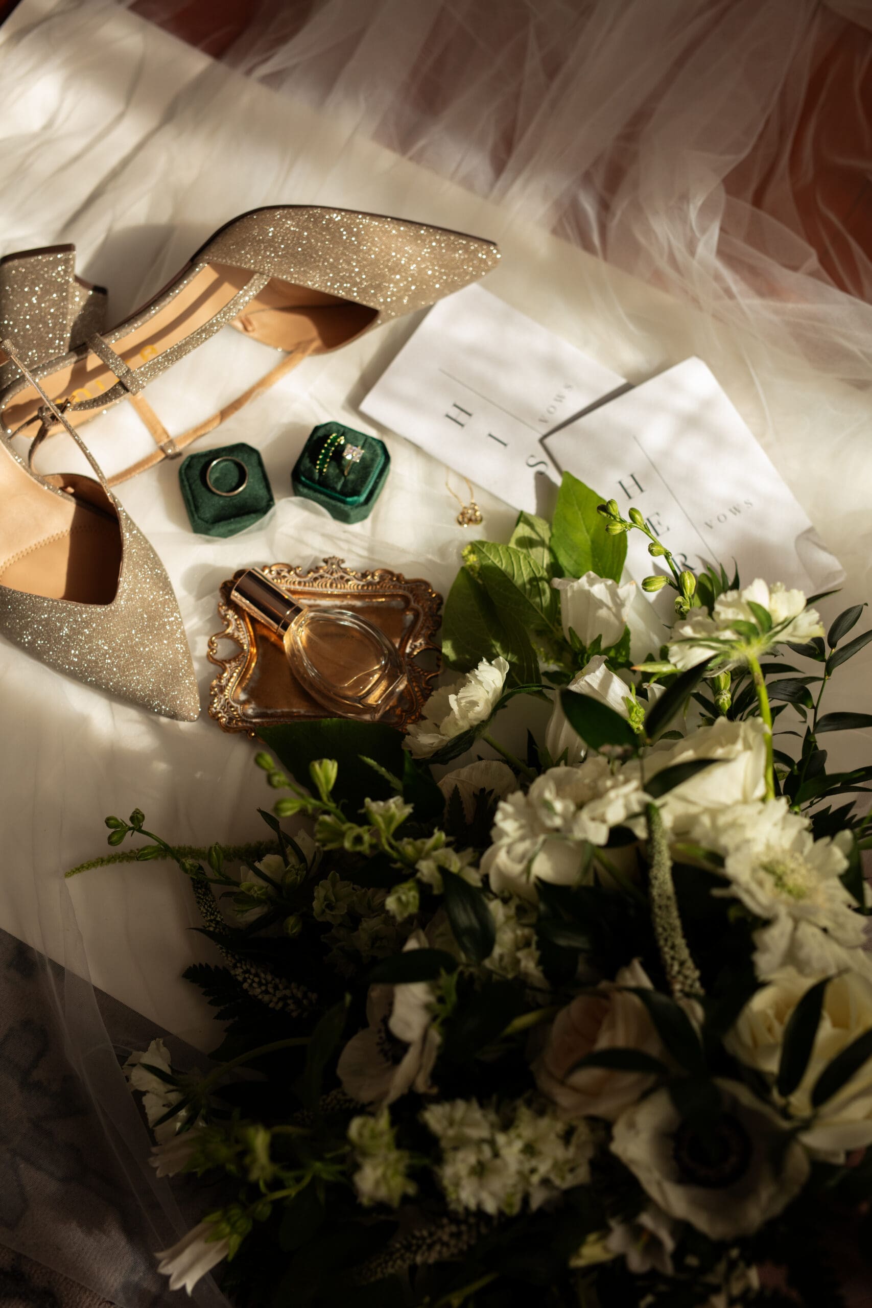 Thoughtful flat lay featuring glittering heels, heirloom rings, vow booklets, and a white floral bouquet in warm natural light.