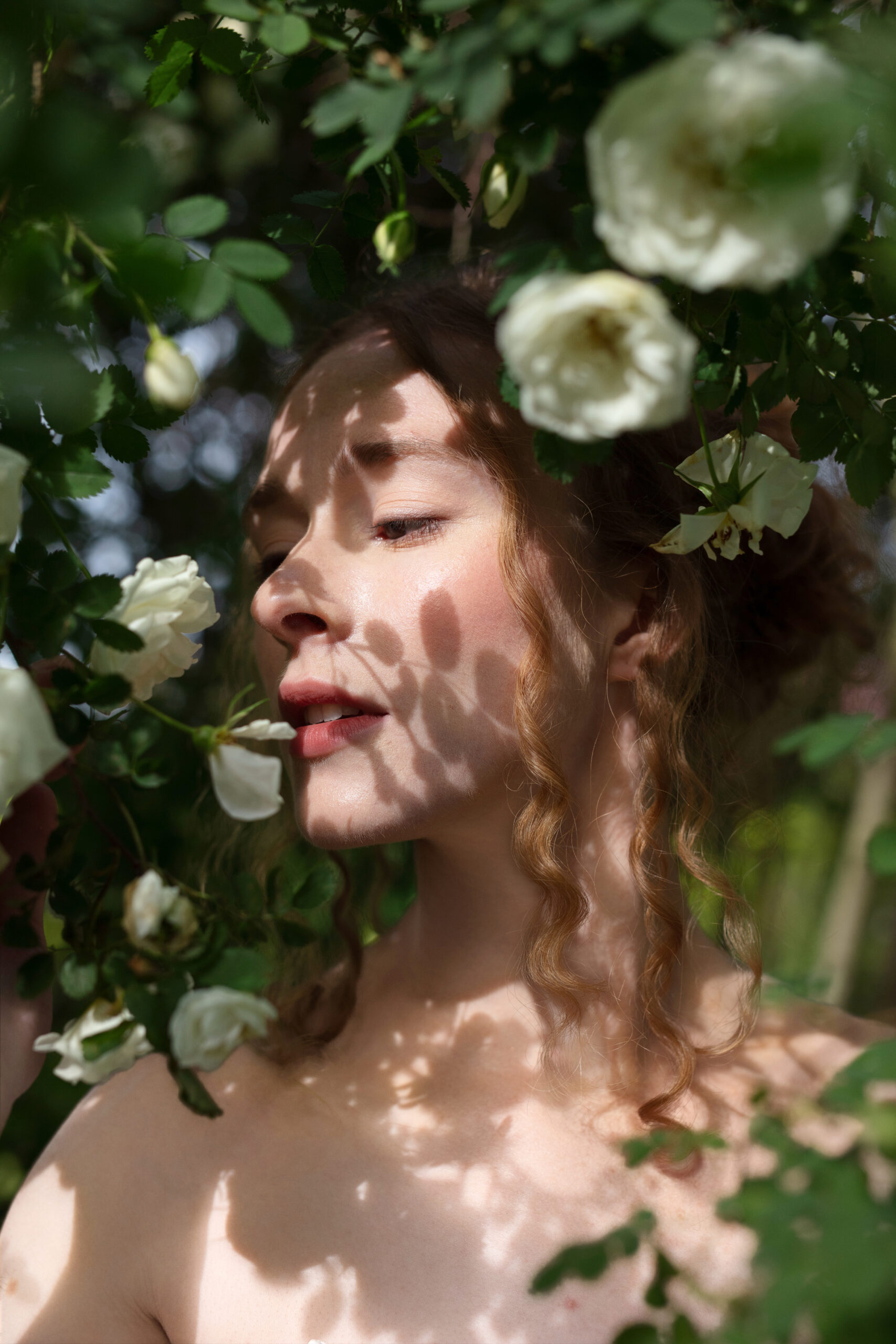 A bride stands in dappled sunlight beneath white garden roses, the shadows playing softly across her face and skin.