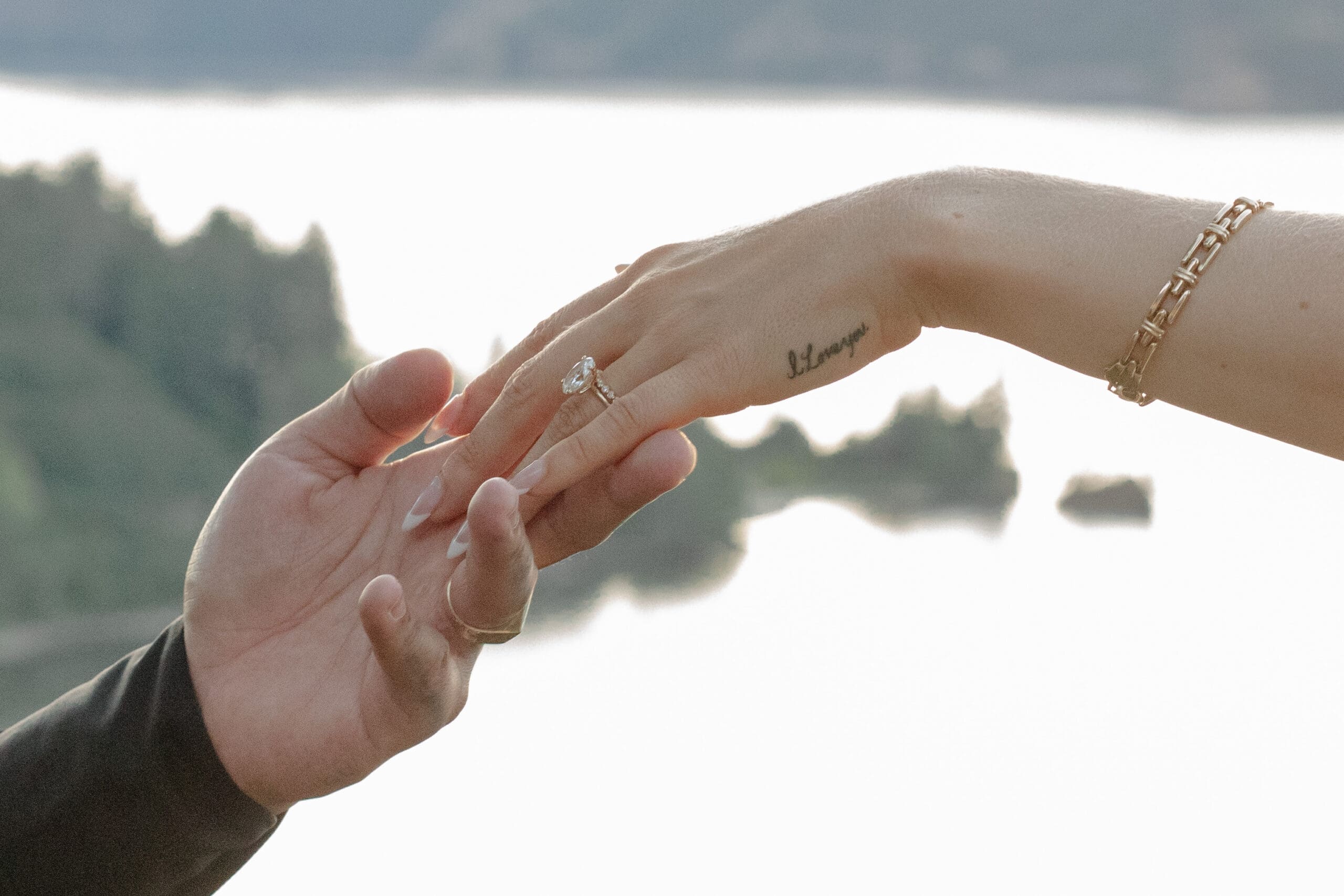 Close-up of two hands reaching for each other, a soft and emotional moment with a mountain lake backdrop and a delicate engagement ring in view.