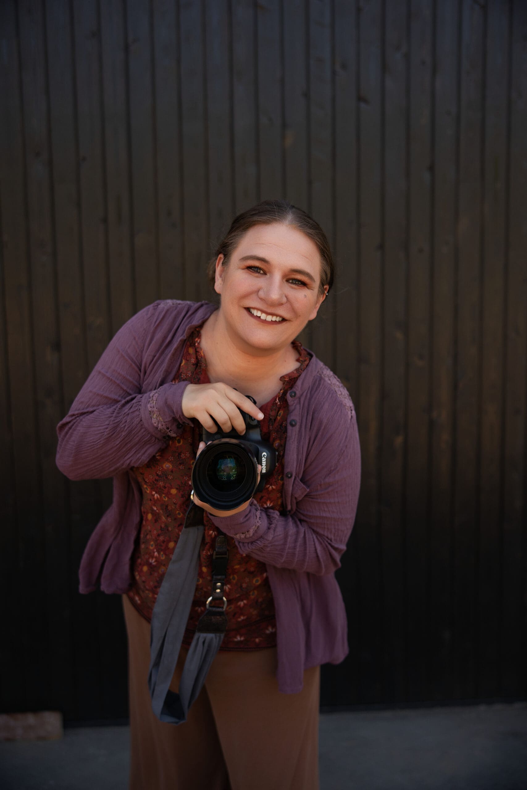 Washington wedding photographer smiling while holding a camera, standing confidently against a dark wooden wall.
