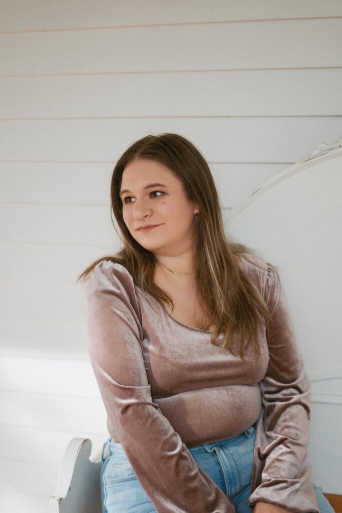 Soft natural light falls across a woman in a rose velvet top, seated against a white paneled wall in a quiet, reflective moment.