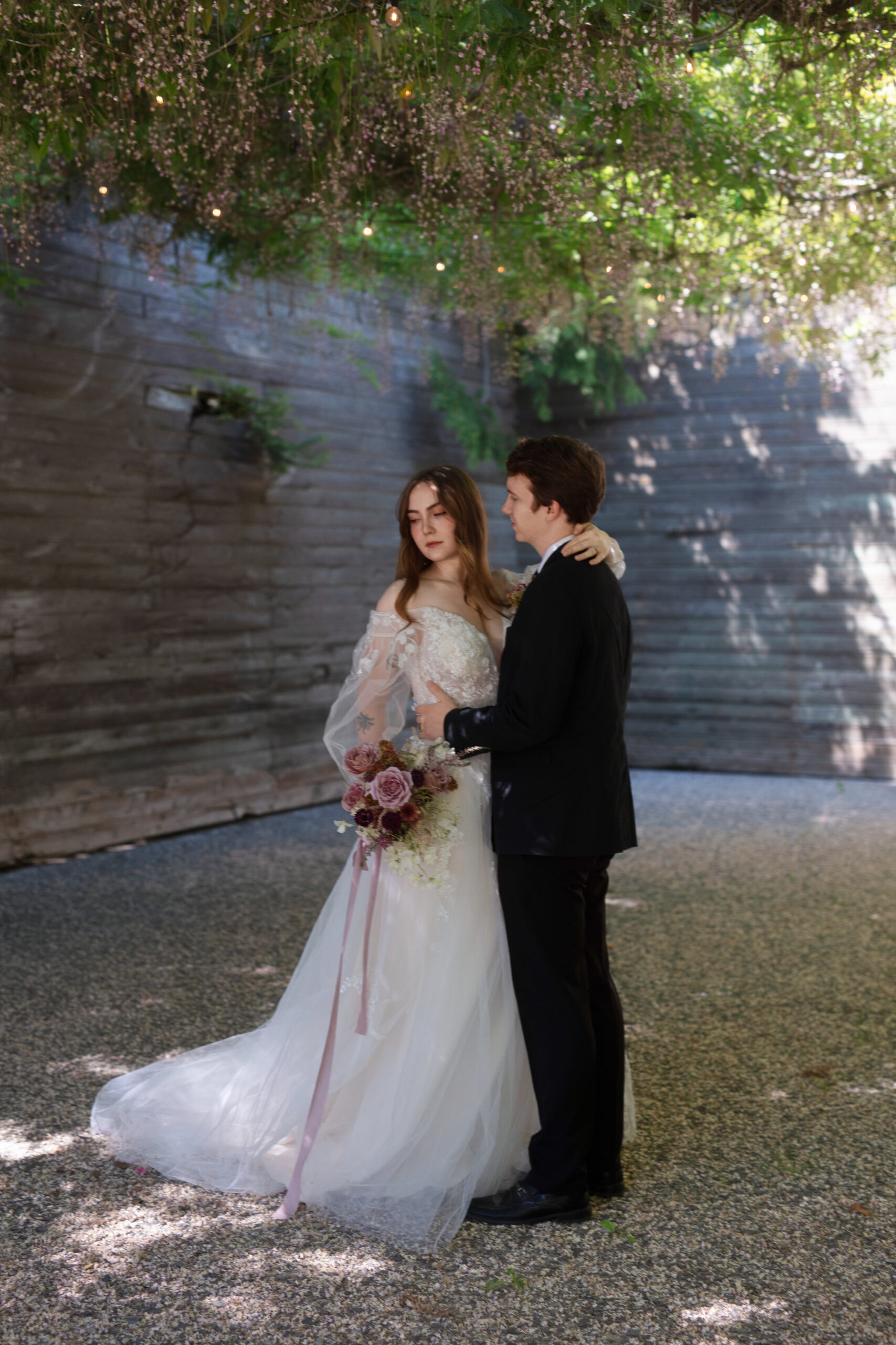 Bride and groom sharing a romantic moment beneath flowering vines and string lights.