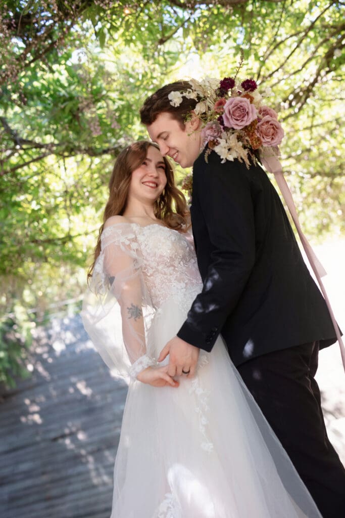 Bride and groom smiling under lush greenery during their wedding day, holding hands and enjoying a romantic moment together.
