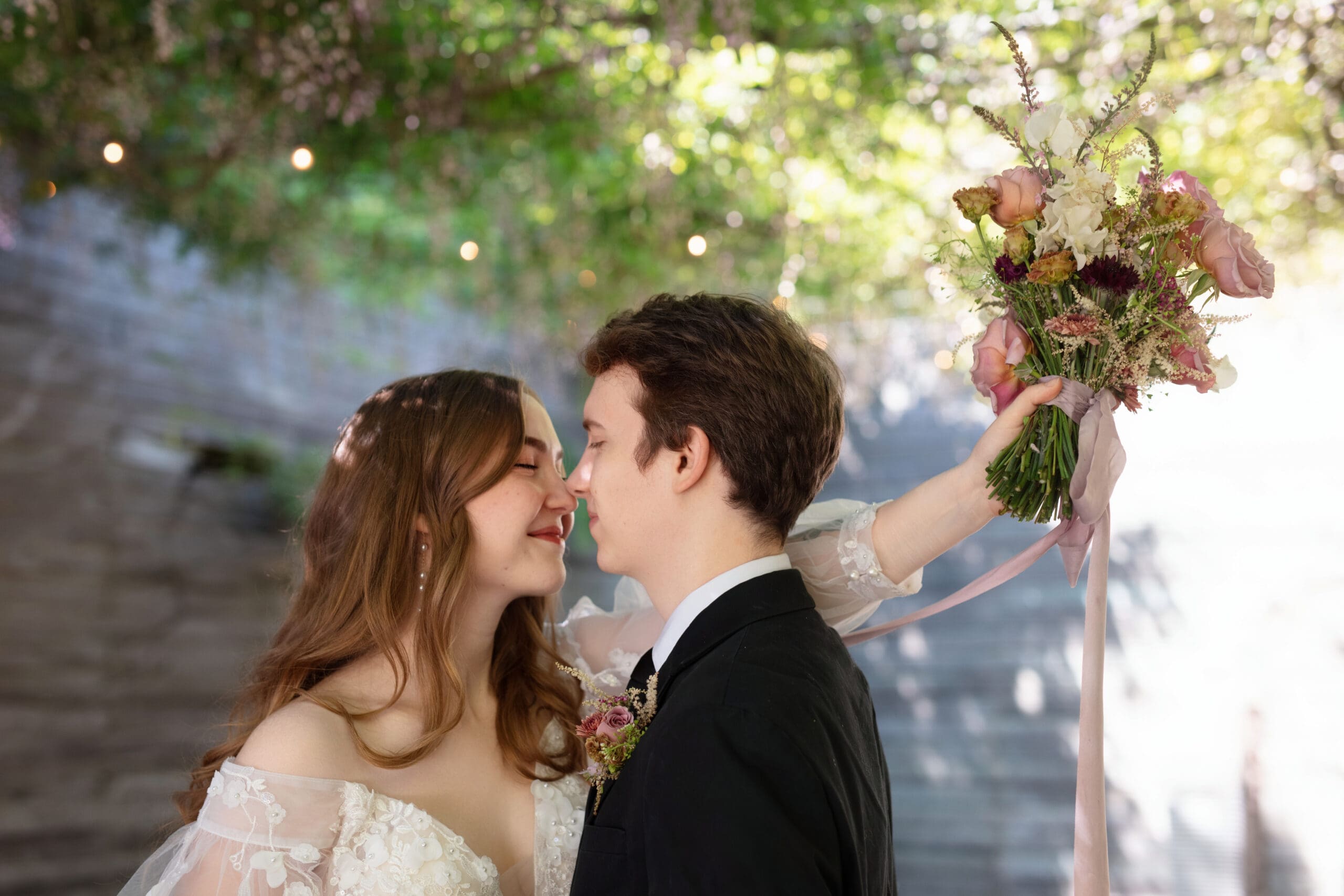 Bride and groom touching noses while bride holds pink bridal bouquet behind groom's head