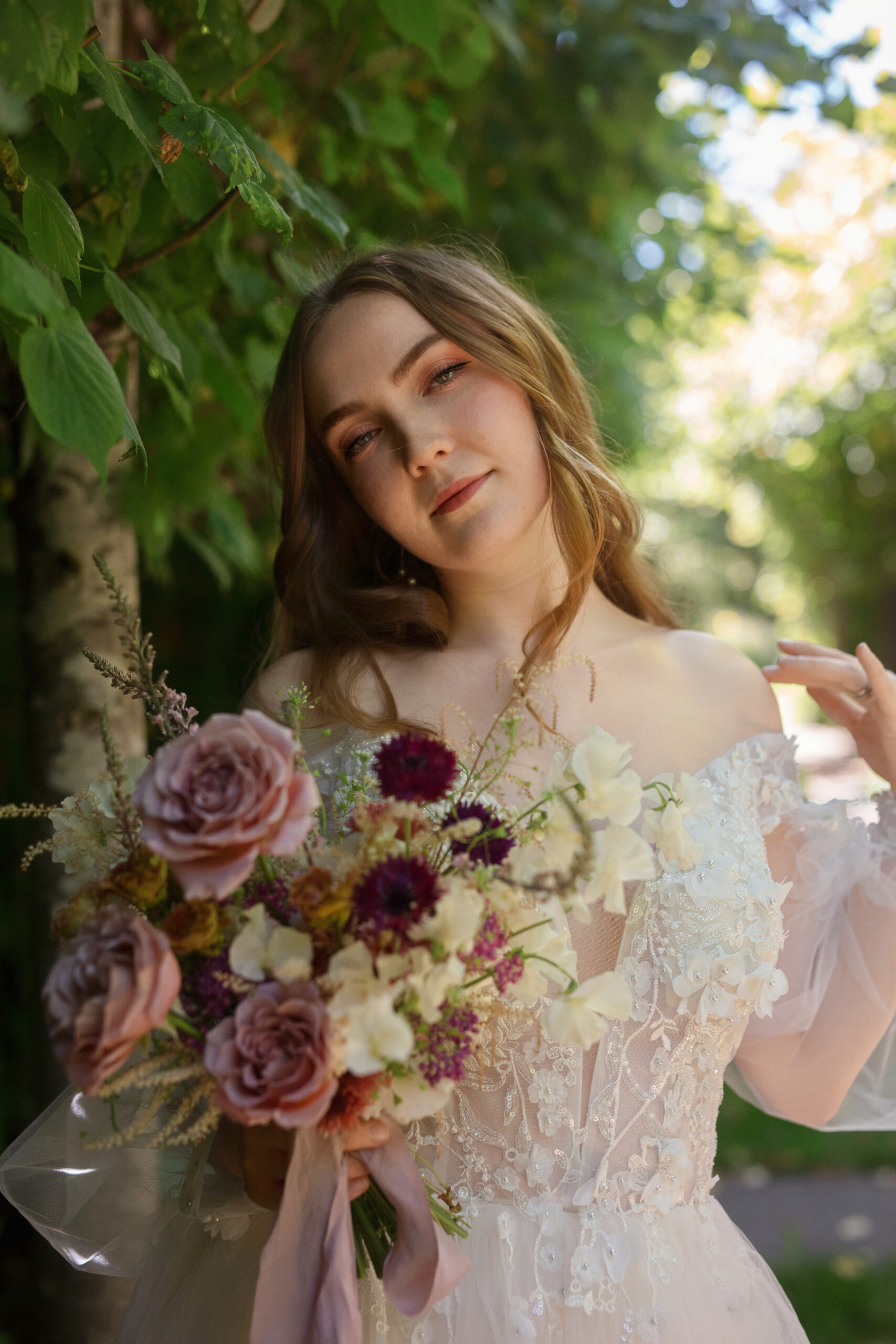 Bride holding a pastel-toned bouquet in front of trees and filtered sunlight.