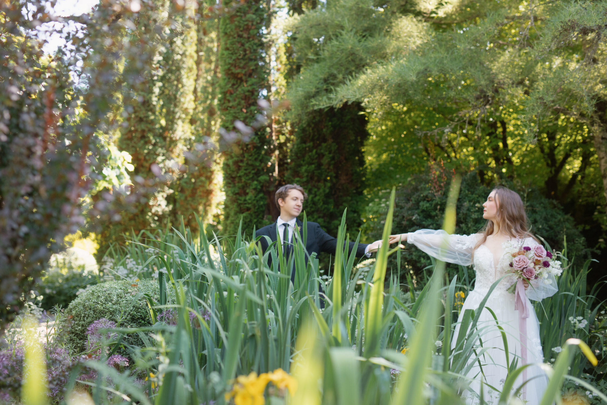 Groom reaching for the bride’s hand as she leads him through tall greenery, bouquet in hand at Briarwood Estate