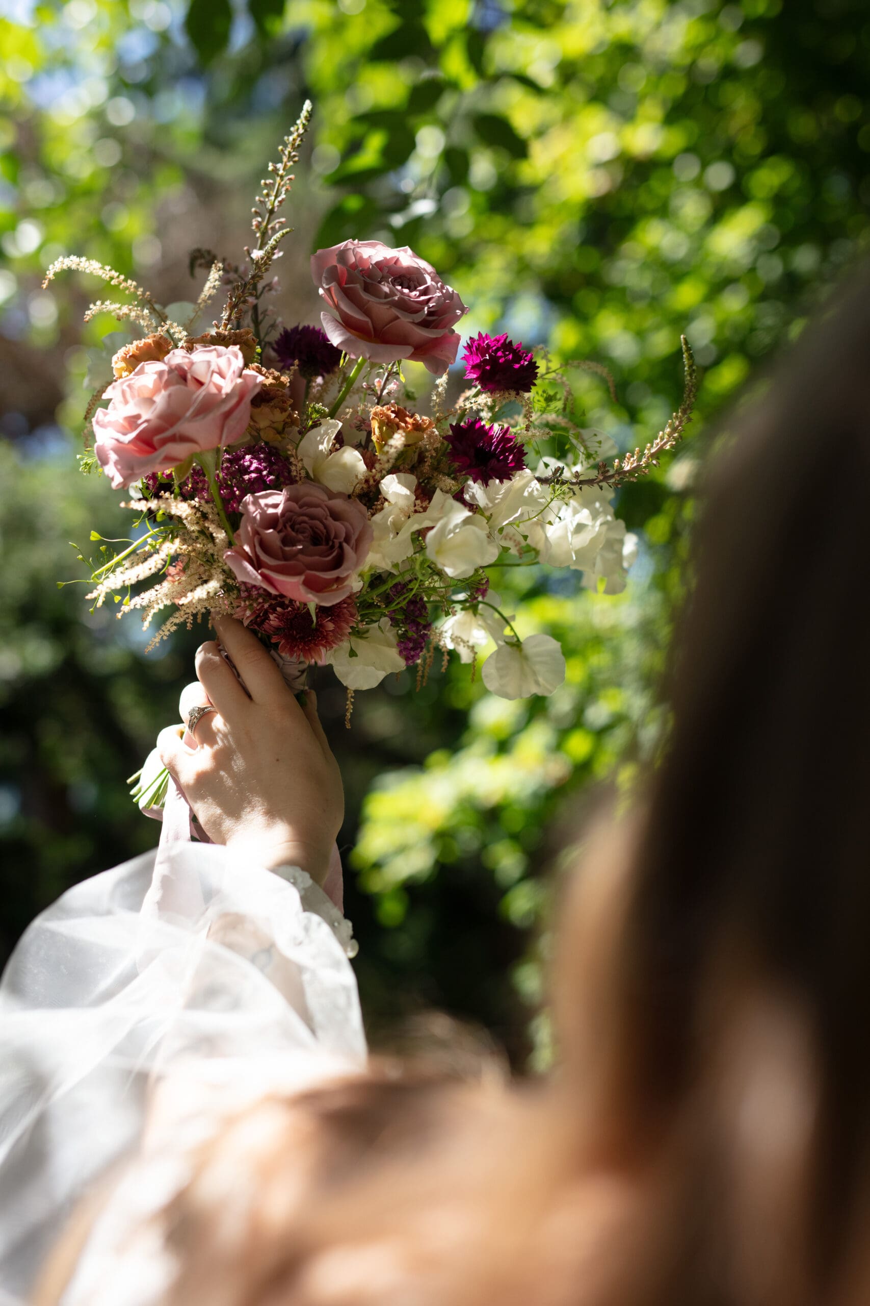 Bride raising bouquet of pink and white flowers under filtered garden light.