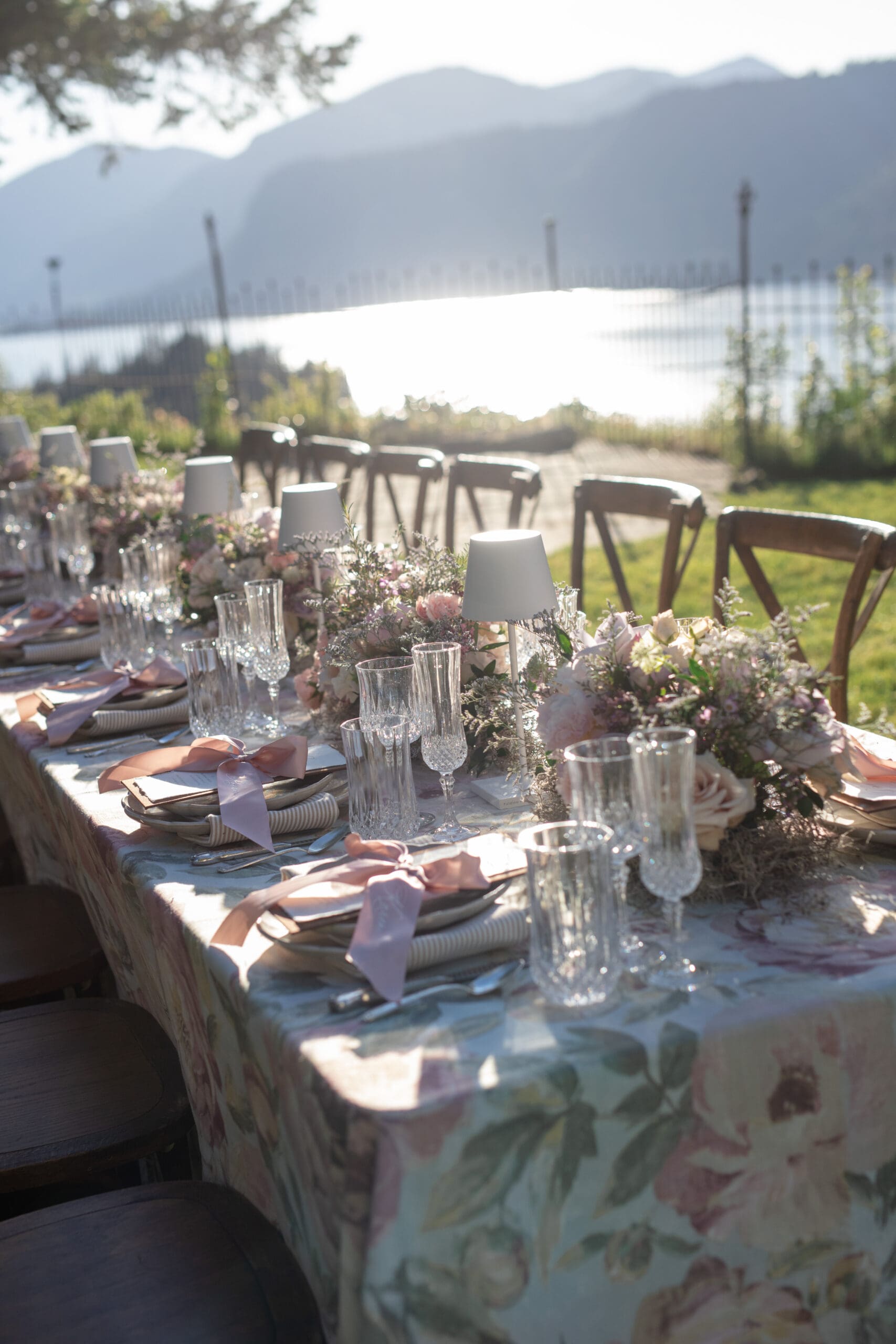 An elegant outdoor reception table is set with pink and white floral arrangements, crystal glassware, and blush napkins on a floral tablecloth, surrounded by wooden chairs beneath towering evergreens.
