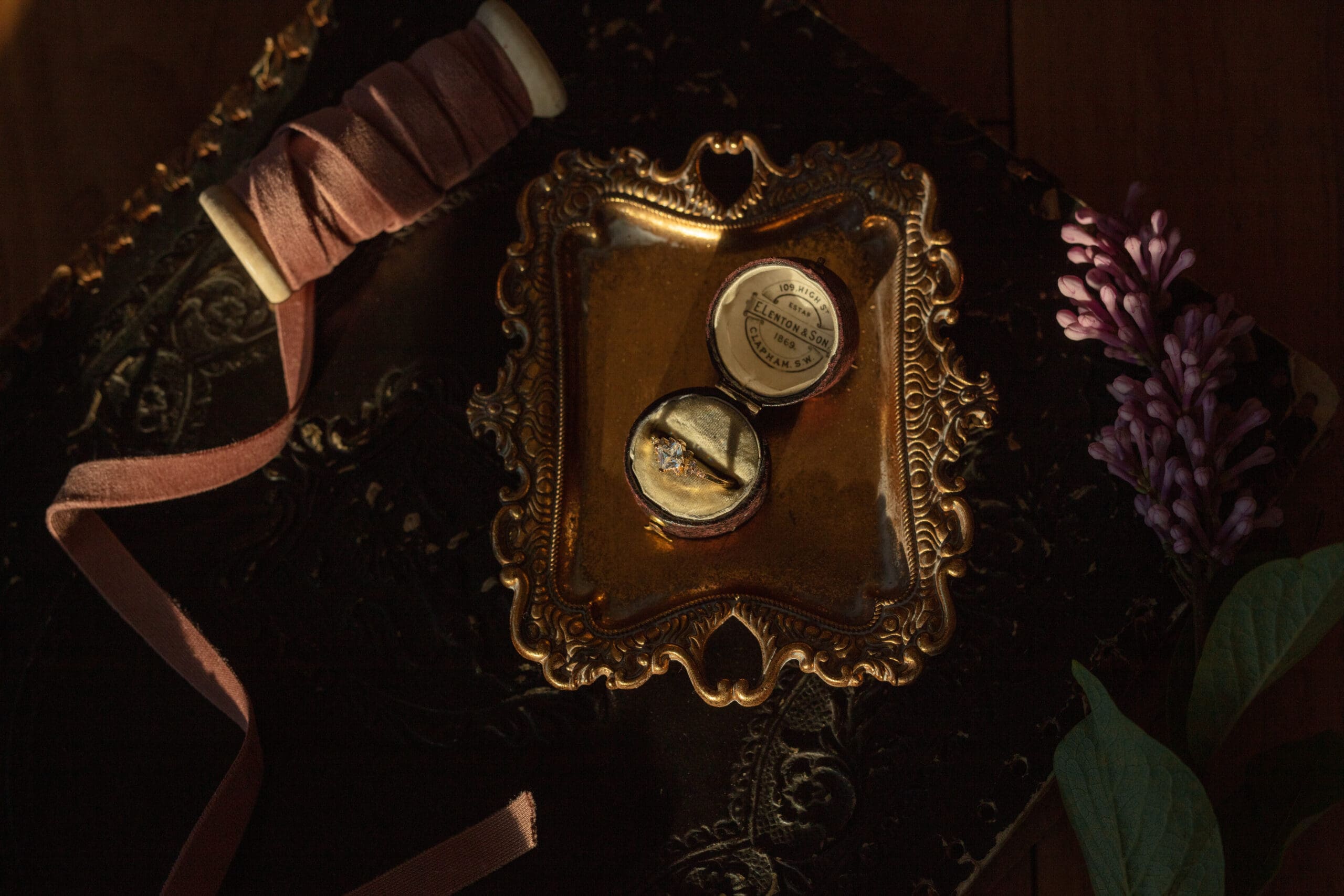 A vintage ring box with an engagement ring rests inside an ornate gold tray, surrounded by velvet ribbon, lilac flowers, and textured black fabric.