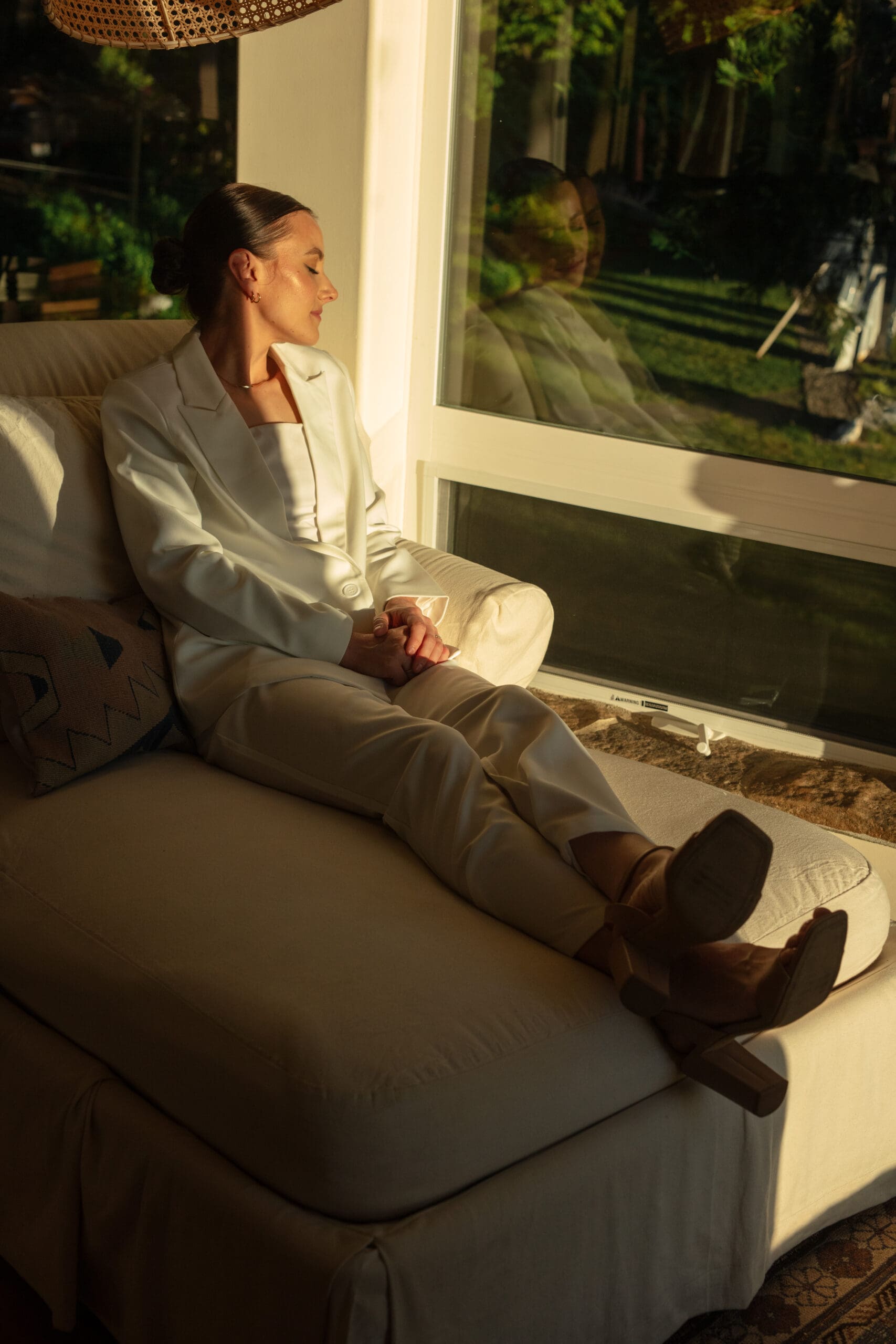A woman in a white suit lounges peacefully on a cream chaise near a window, bathed in golden hour sunlight, with her reflection visible in the glass.