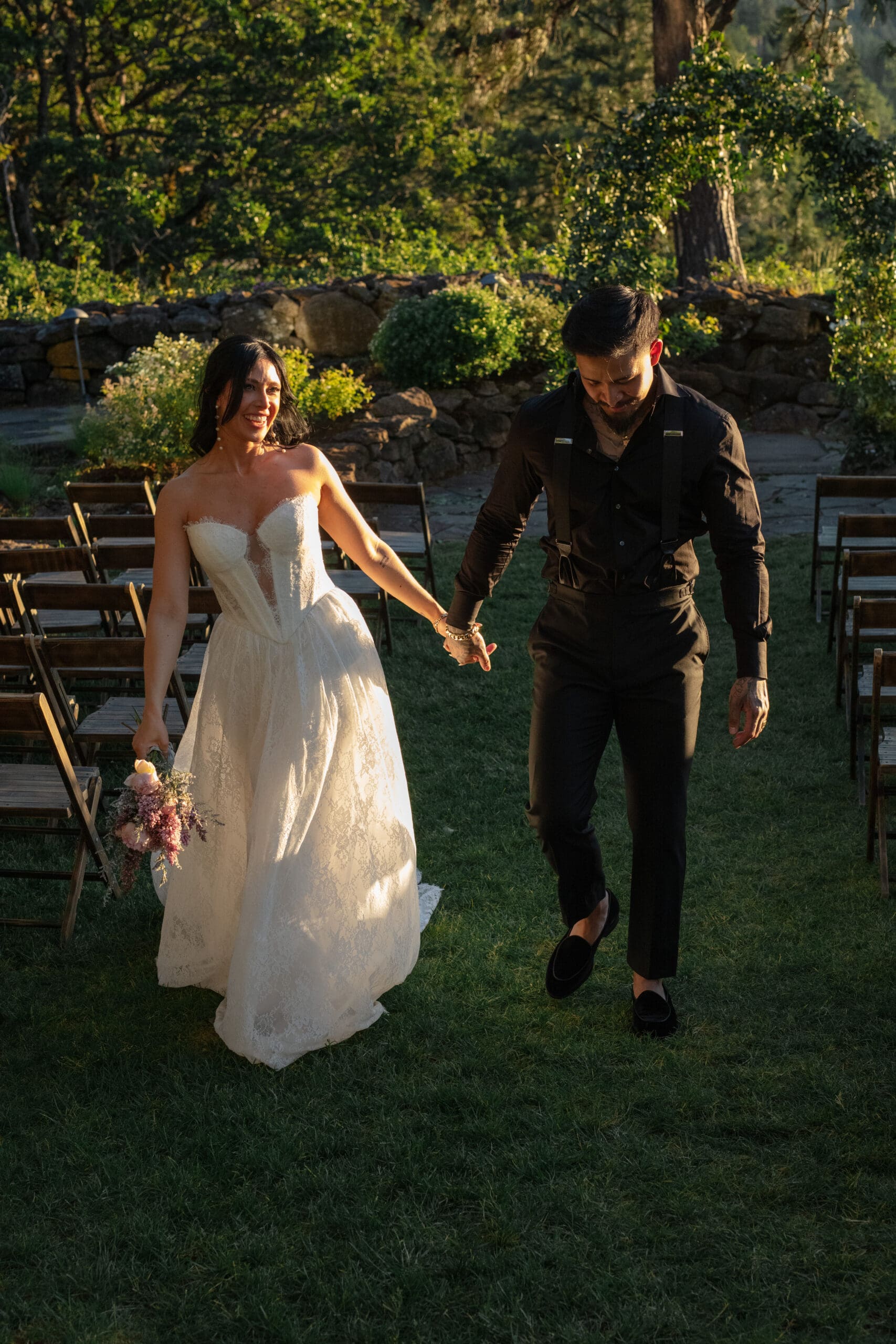 Newlyweds walk hand-in-hand down an outdoor aisle lined with wooden chairs, smiling and glowing in the soft golden light of sunset.

