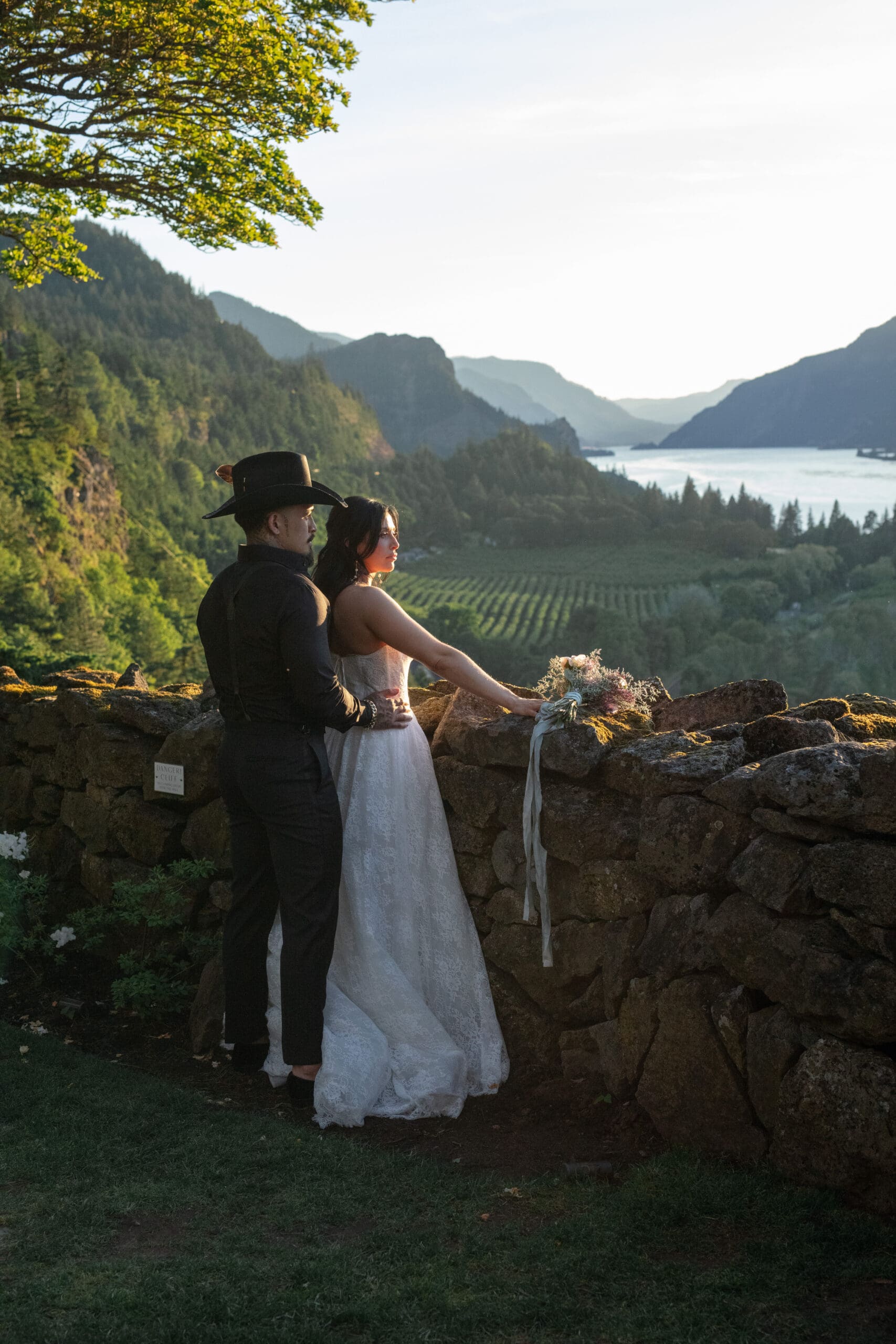 A couple stands together overlooking a stunning valley and river at golden hour, the groom in a black cowboy hat and the bride in a white lace gown with her bouquet resting on a stone wall.