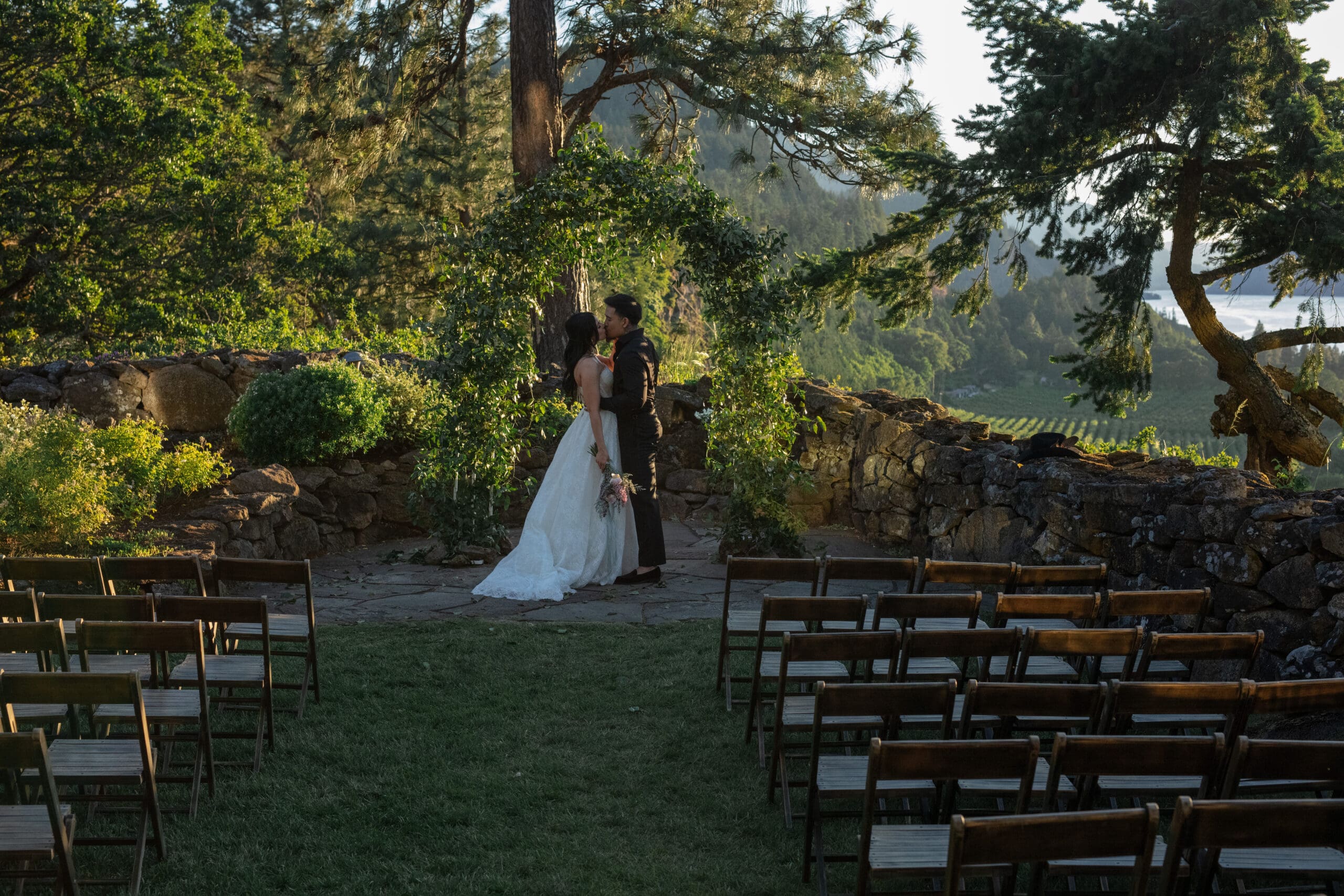 A couple shares a kiss under a leafy green arch at their outdoor wedding ceremony, framed by a stone wall and overlooking a lush mountain valley in the golden glow of sunset.

