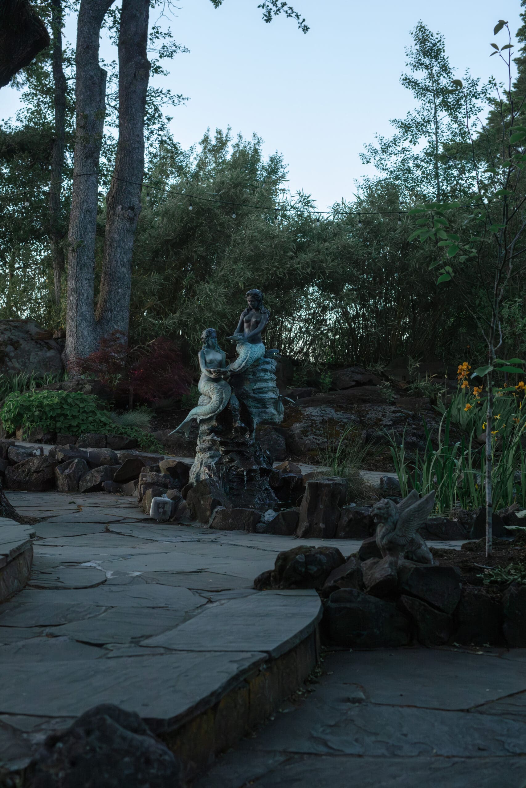 A whimsical garden statue of two mermaids sits on a stone base, surrounded by lush greenery, tall trees, and a paved stone path in a peaceful forested area.

