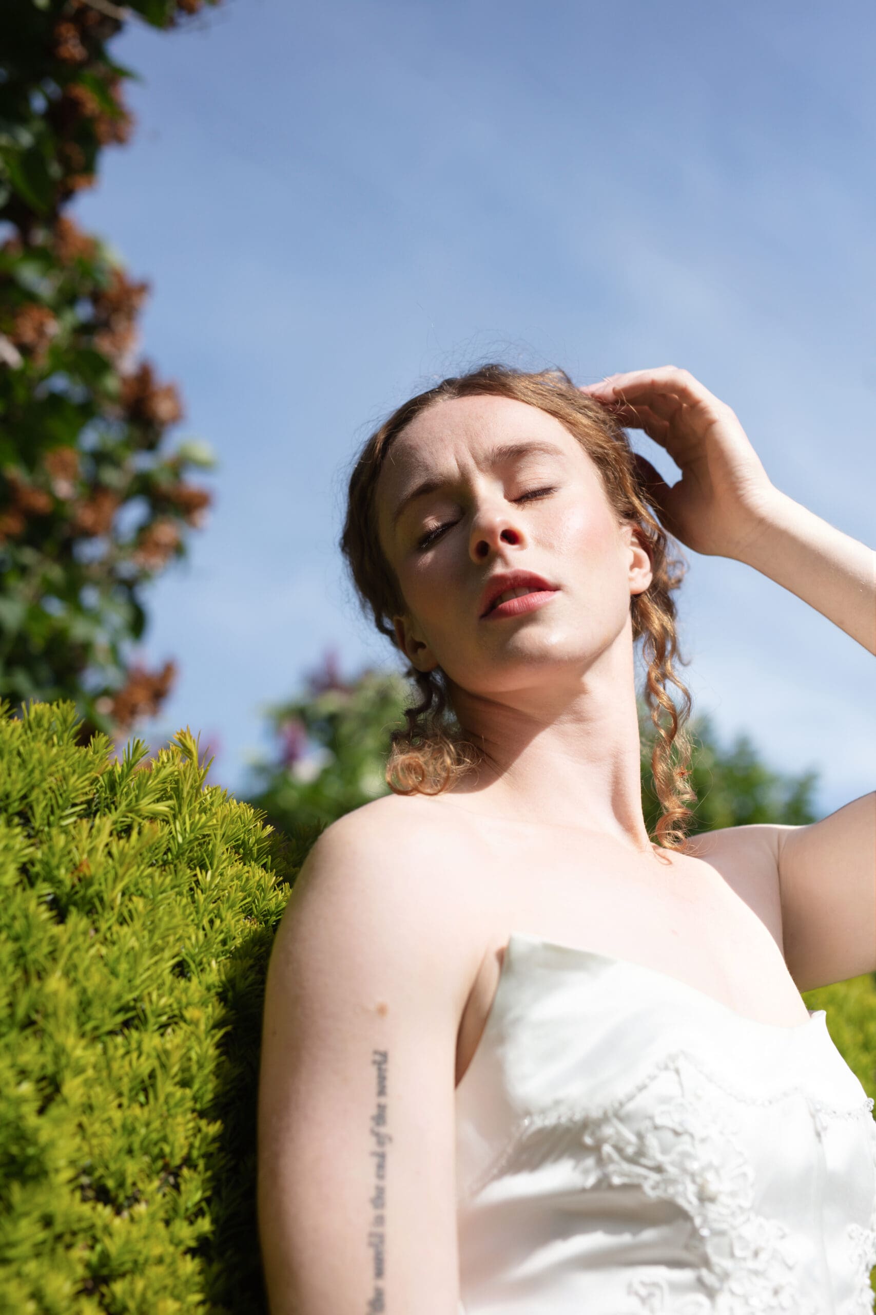 Bride in soft sunlight with eyes closed and hand in hair, standing against a green hedge at Christianson’s Nursery wedding