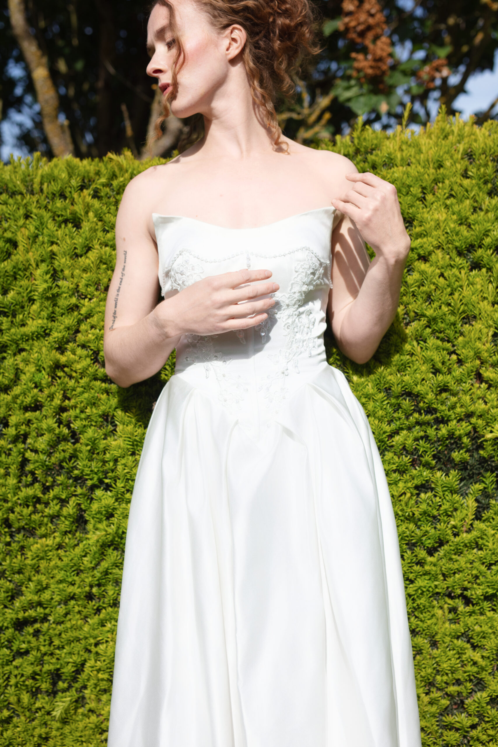 Detail shot of vintage wedding dress and bride's tattooed arm in front of vibrant hedges.
