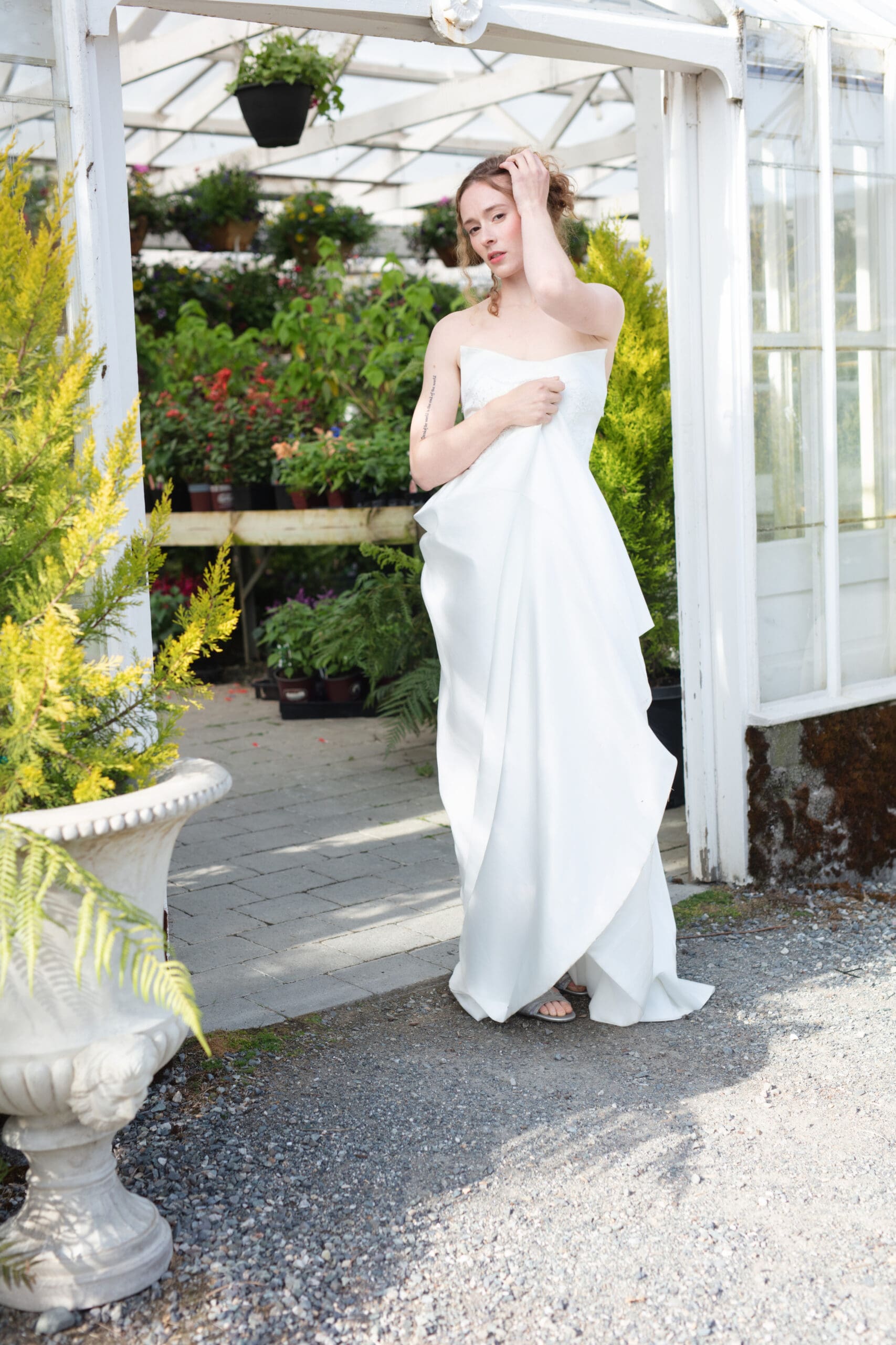 Bride standing in the greenhouse doorway wrapped in her gown, framed by ferns and blooming potted plants.
