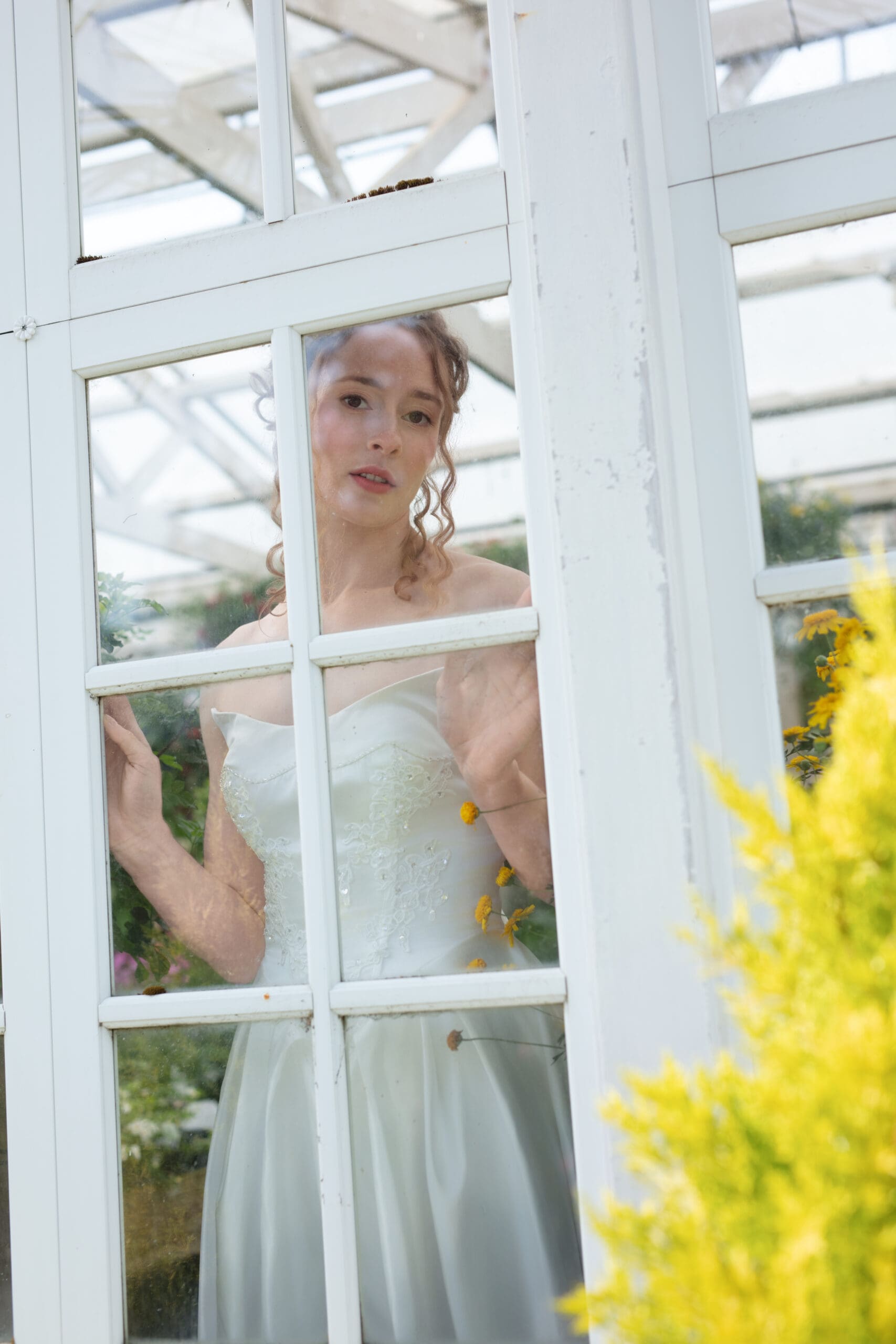 Artistic bridal portrait behind greenhouse glass, highlighting soft curls and painterly natural light.