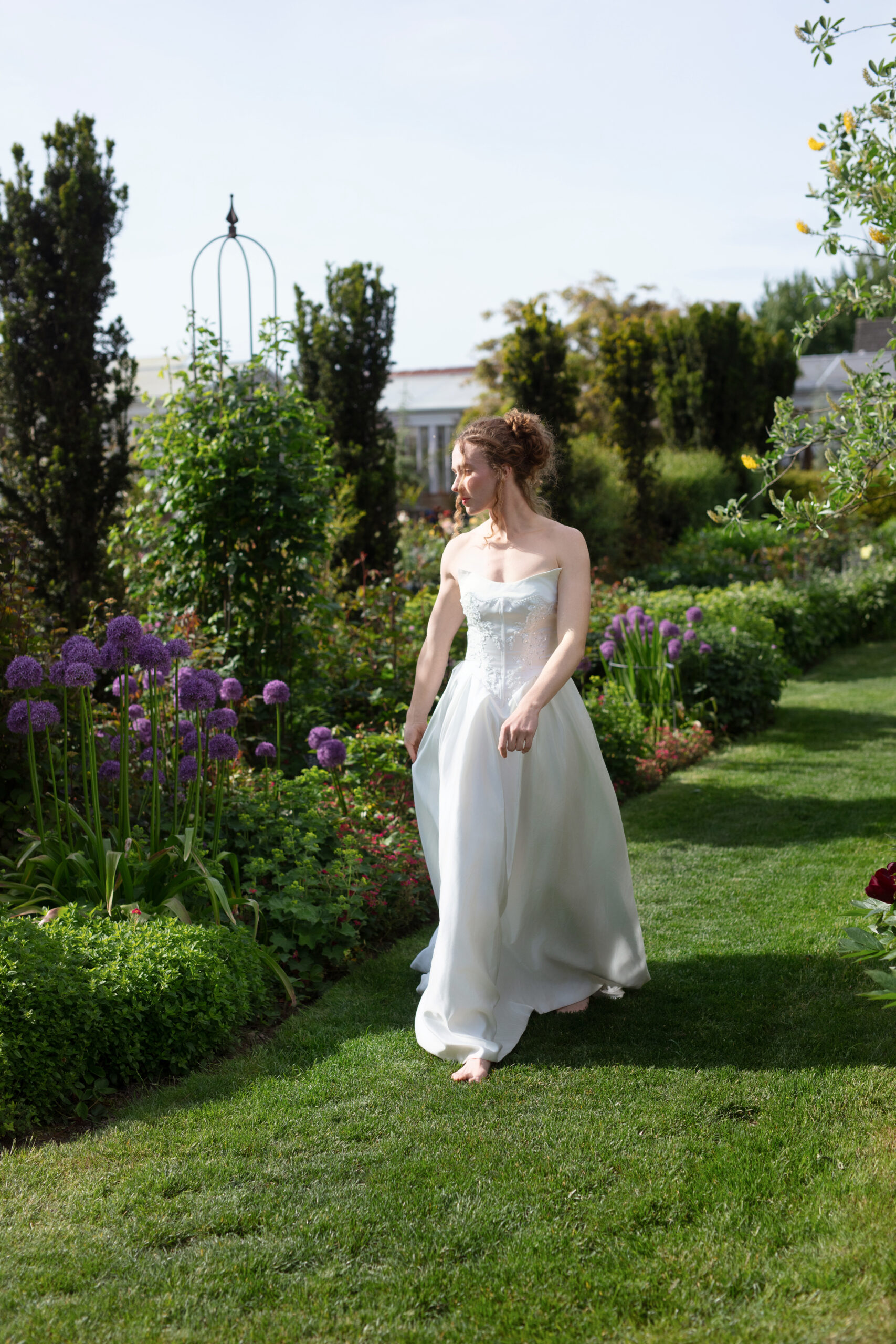 Bride walking along manicured garden path lined with purple alliums and trimmed hedges at Christianson’s Nursery wedding.