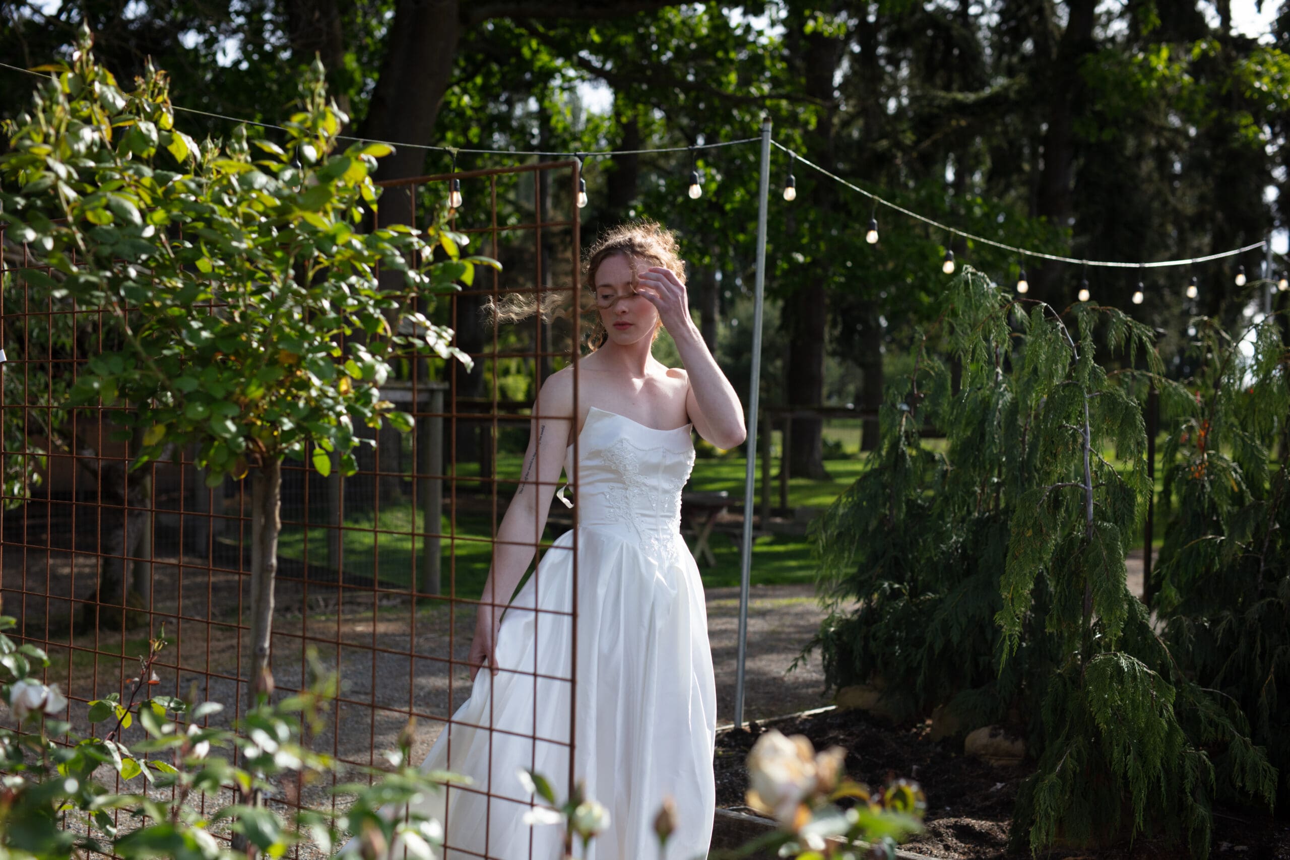 Candid bridal moment in the garden as wind lifts her hair, surrounded by string lights and trees.