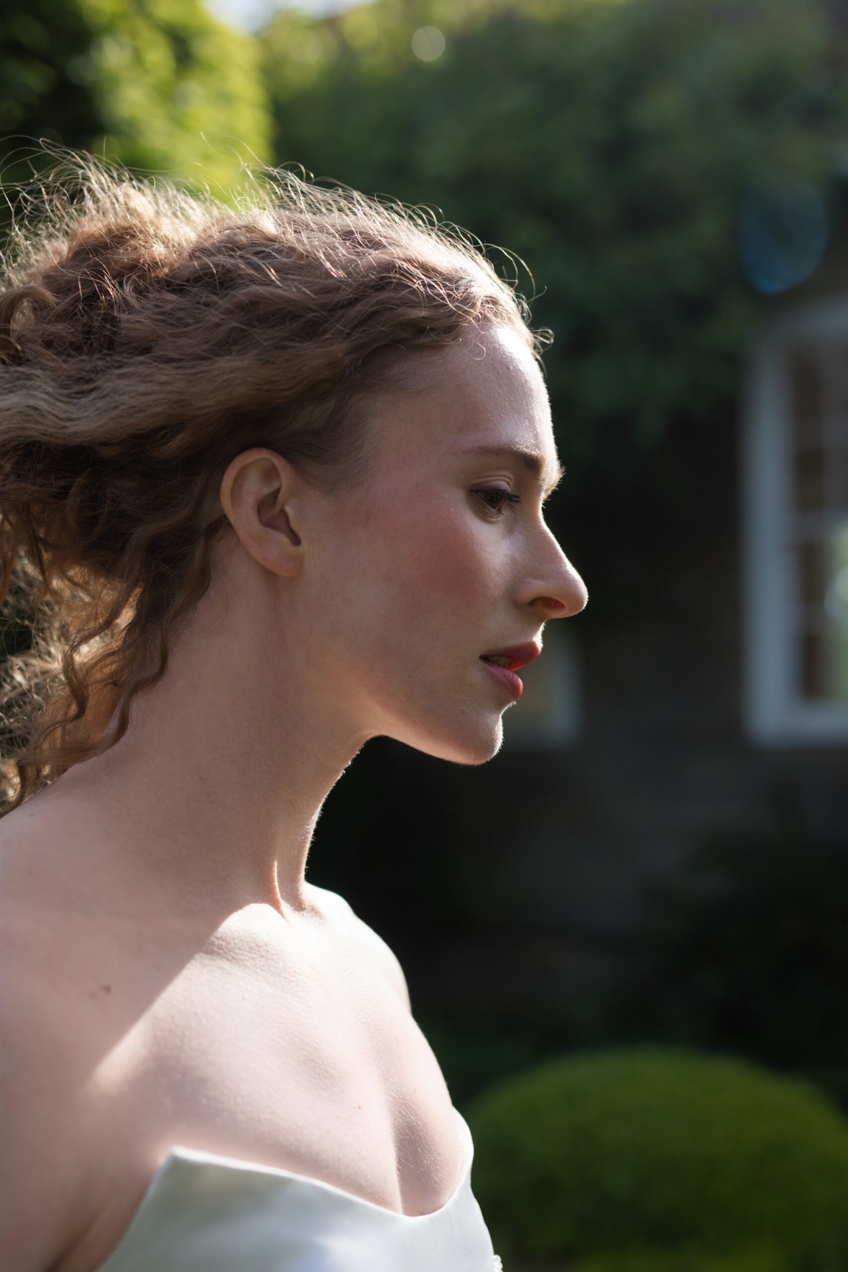 Candid bridal profile portrait in warm afternoon light with greenery and a cottage in the background.