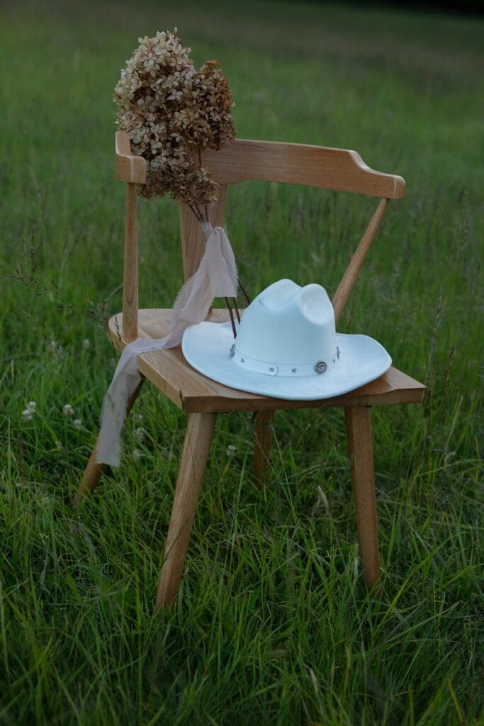 A white cowboy hat and dried florals styled on a wooden chair in the grassy field at wind mountain ranch venue.