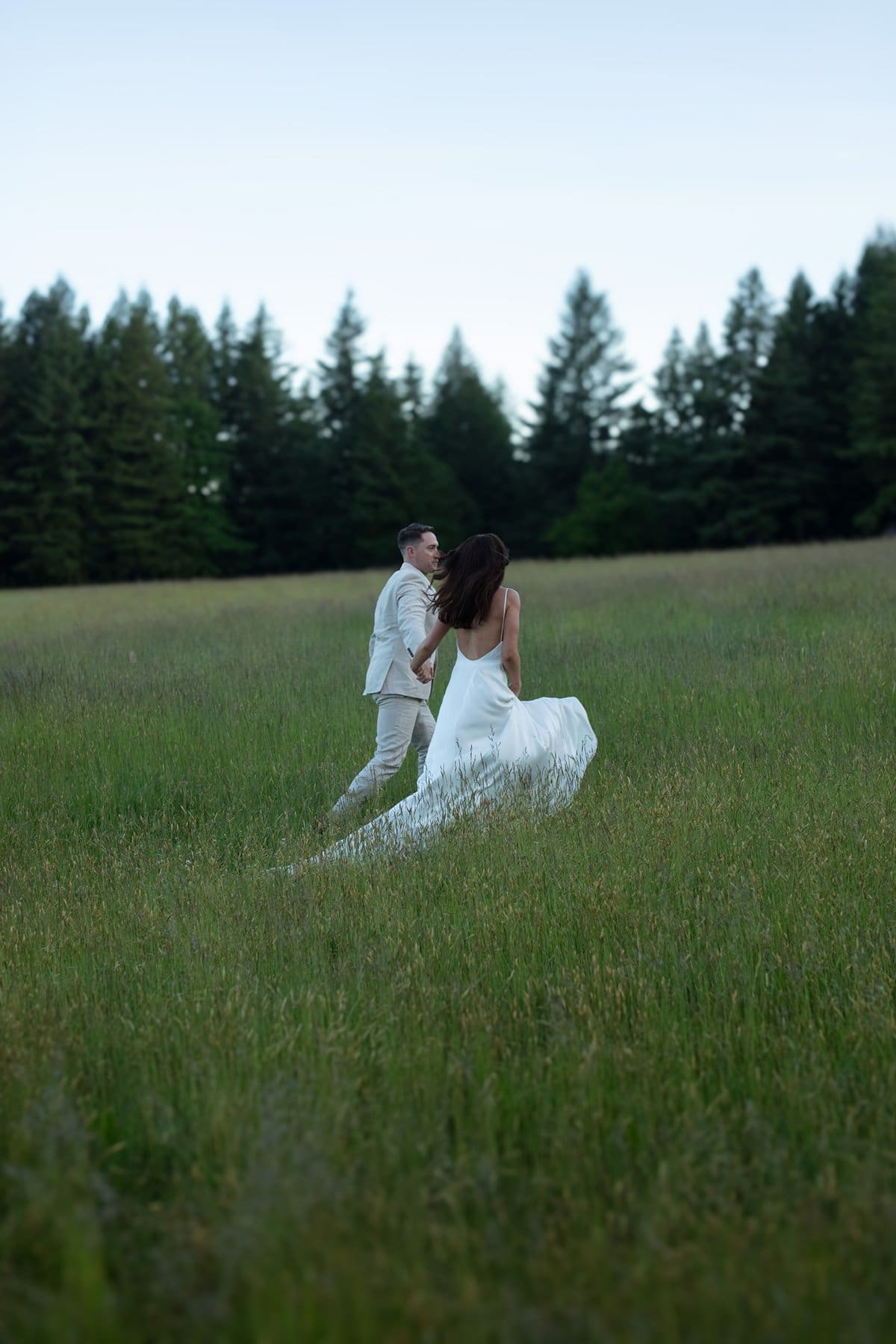Bride and groom run hand-in-hand through a wild grassy field at Wind Mountain Ranch venue, framed by forest and open sky.