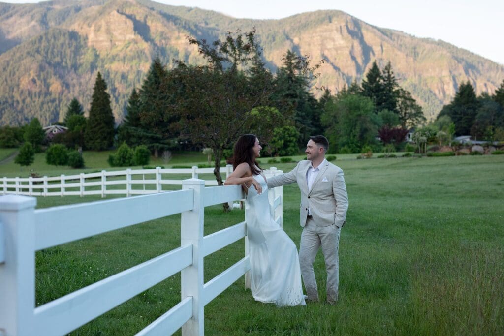 Bride leaning on a white fence smiling at her groom during golden hour in the mountains