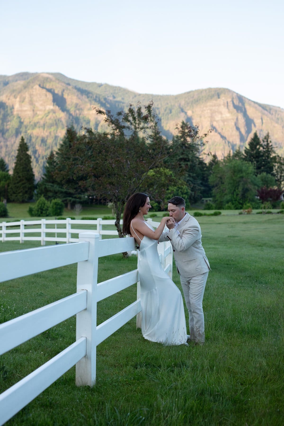Romantic sunset moment as a couple holds hands beside a white fence with mountain views in the background.