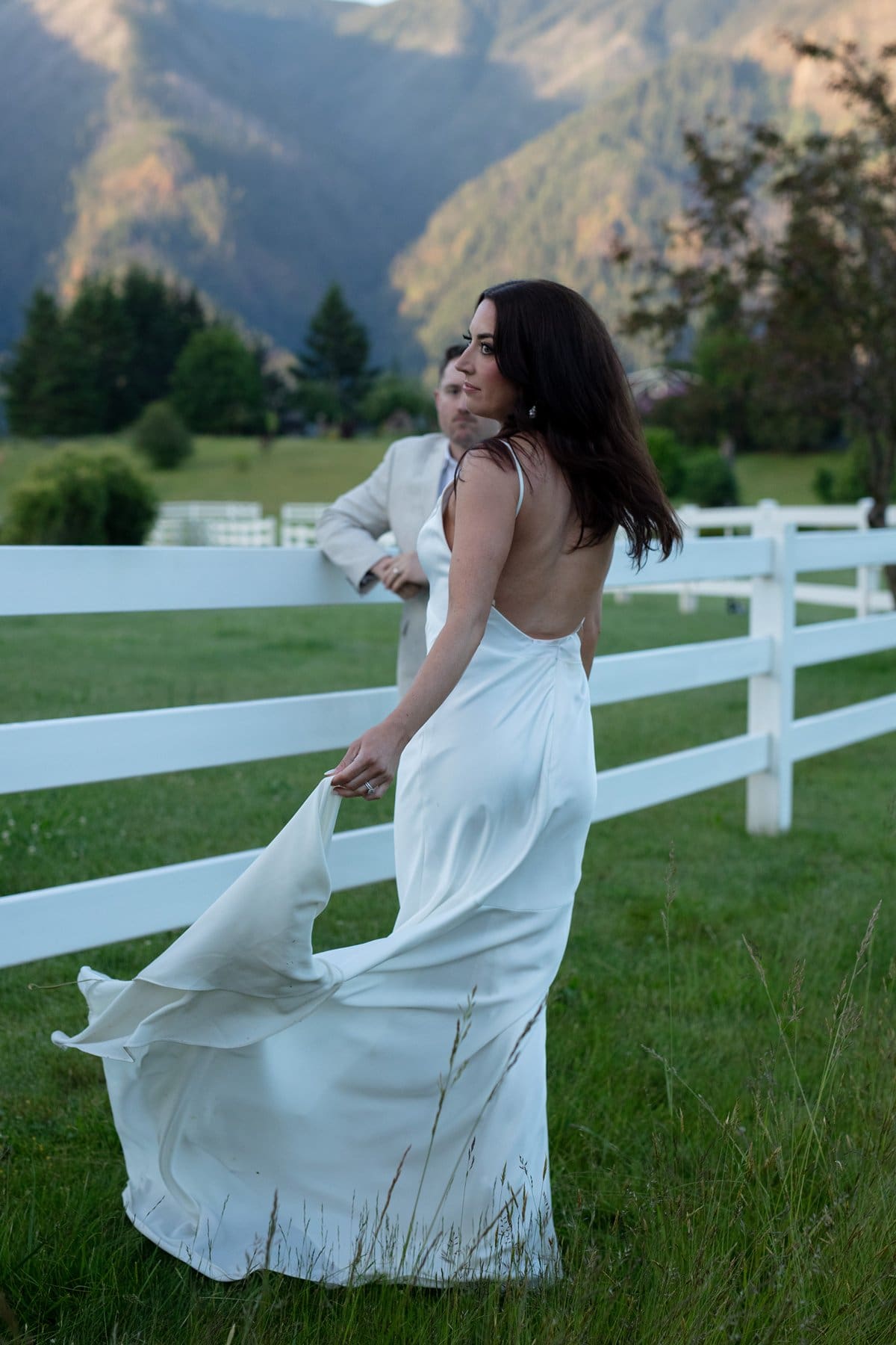 Bride in a satin gown looks over her shoulder while walking by a white fence at Wind Mountain Ranch venue.