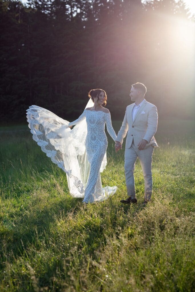 Groom holding bride’s hand as she lifts her wedding train, walking through sunlit grass during their golden hour portraits