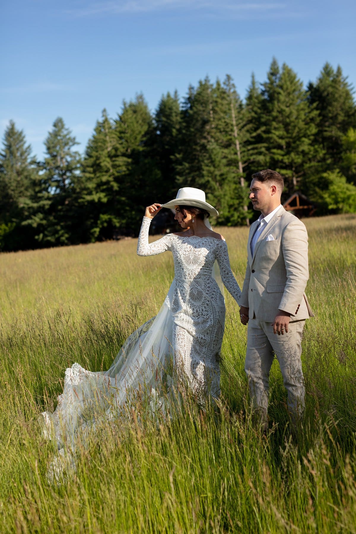 Bride in a lace gown and white hat walks hand-in-hand with her groom through the tall golden grass at wind mountain ranch venue