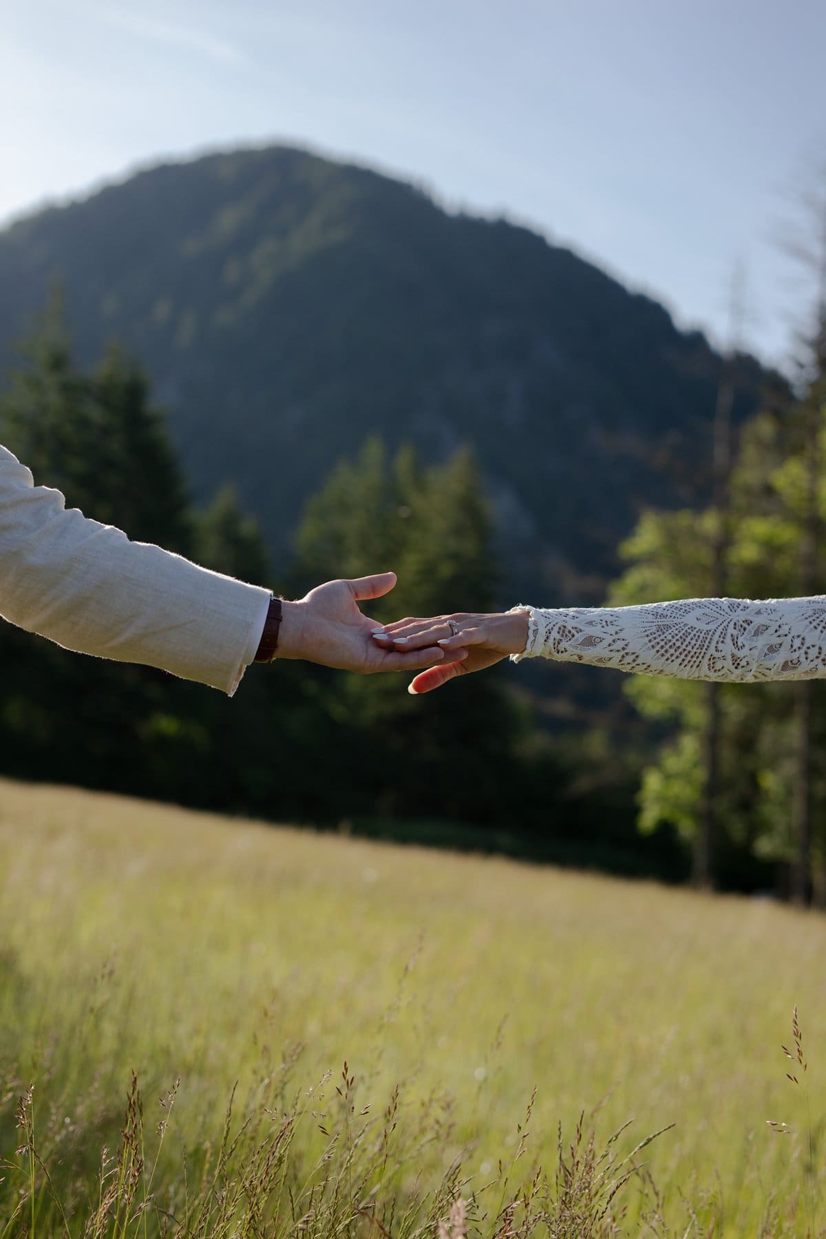 A bride and groom stretch their hands toward each other in a quiet, romantic moment with a mountain backdrop.