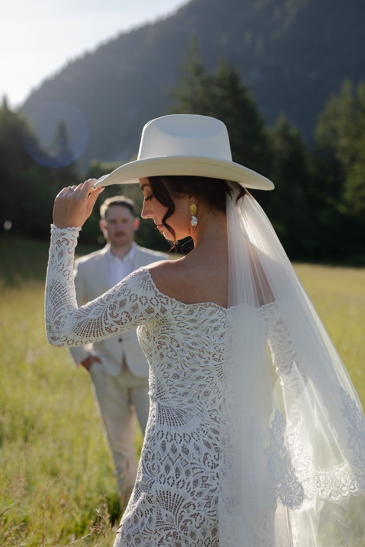 The bride tilts her white cowboy hat as her groom approaches in the grassy field, bathed in soft evening light.