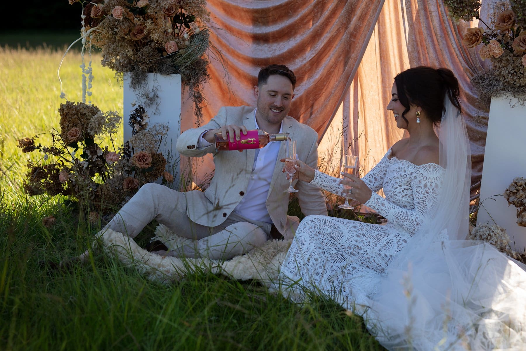 Groom pouring rosé for his bride as they sit on a picnic blanket surrounded by dried florals and a velvet backdrop

