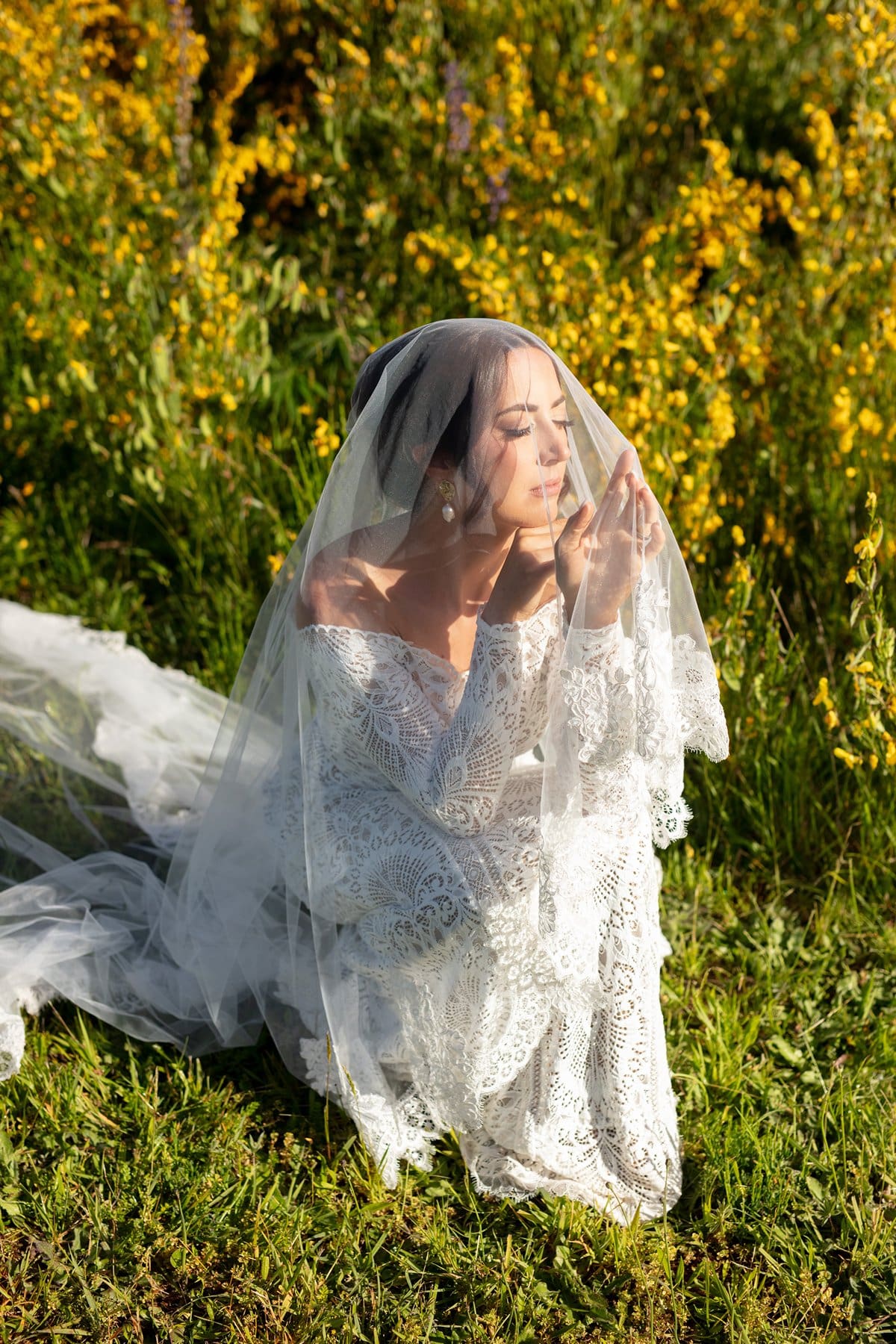 The bride kneels in wildflowers with her veil over her face, hands softly pressed together in the sunlight.