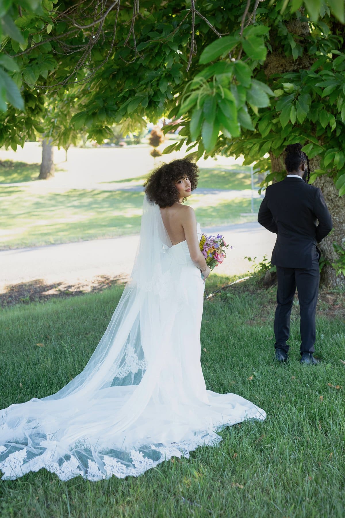 Bride glances over her shoulder with bouquet in hand during a quiet first look moment in the trees.