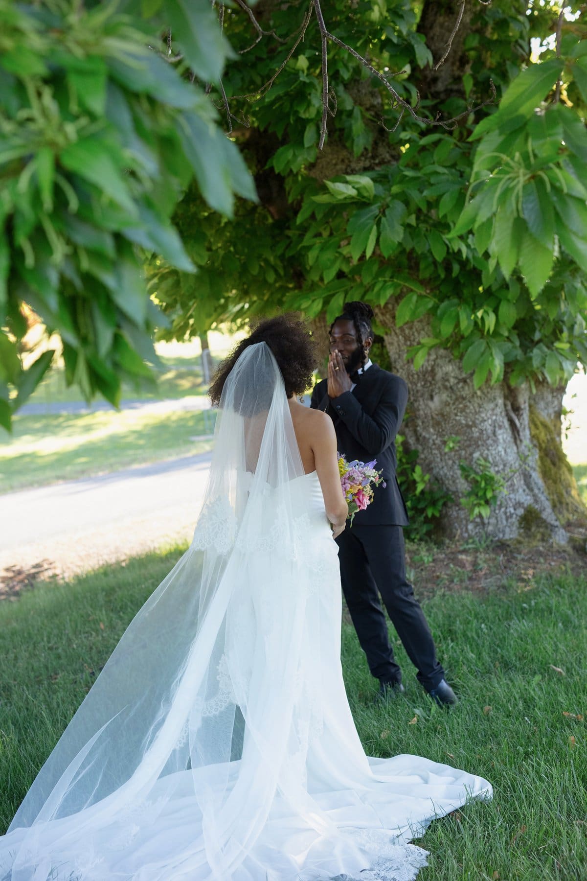 Bride in long veil approaches groom for emotional first look beneath the shade of a large tree.