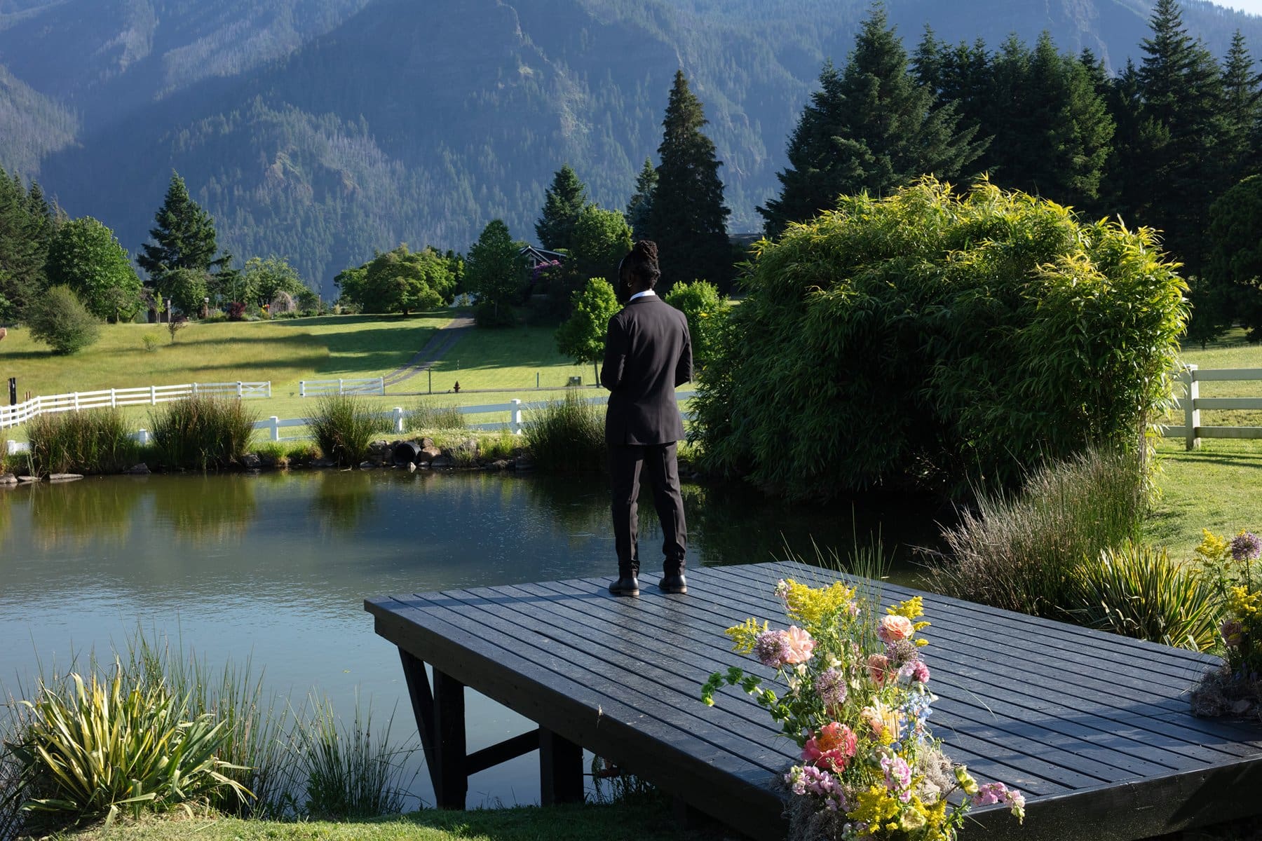 Groom awaits his bride on the dock overlooking the pond and mountain backdrop at wind mountain ranch venue.