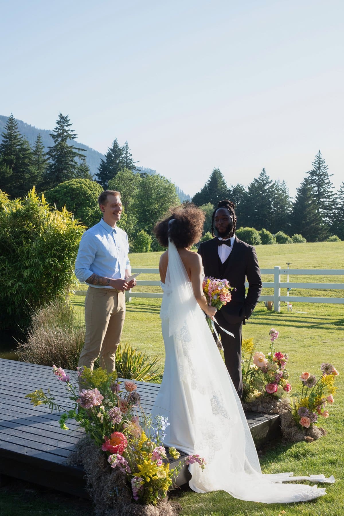 The couple shares vows on the wooden dock ceremony space, surrounded by florals and mountains at Wind Mountain Ranch venue.