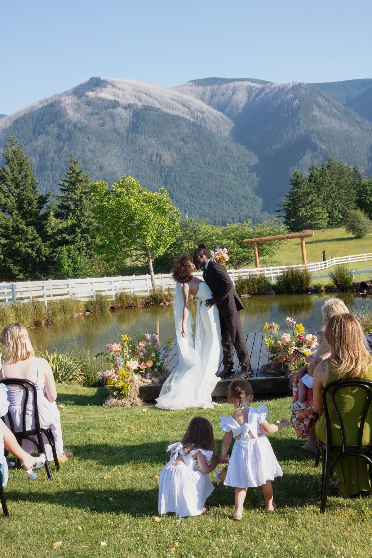 Intimate wedding ceremony by the pond with mountain views, as the bride and groom share a kiss under blue skies.