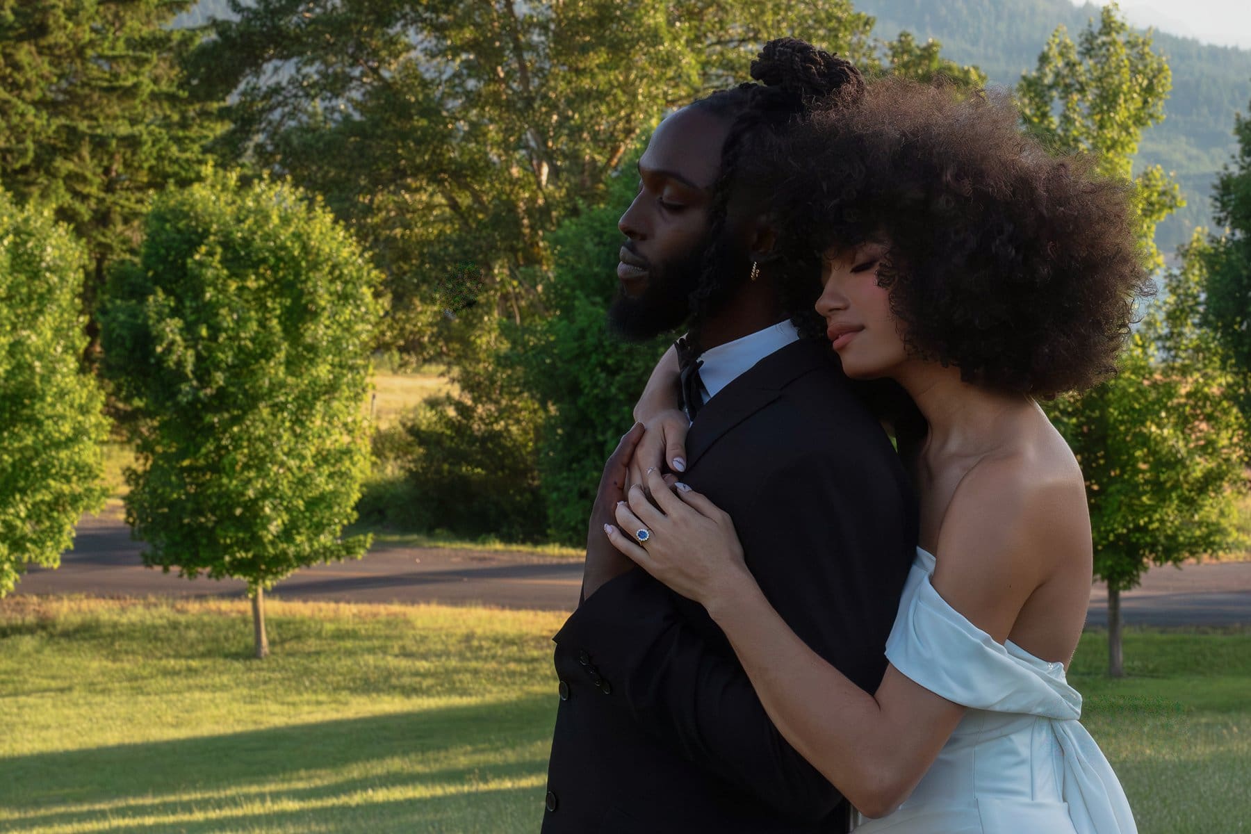 Close-up moment of the bride embracing the groom from behind during golden hour, trees glowing behind them.