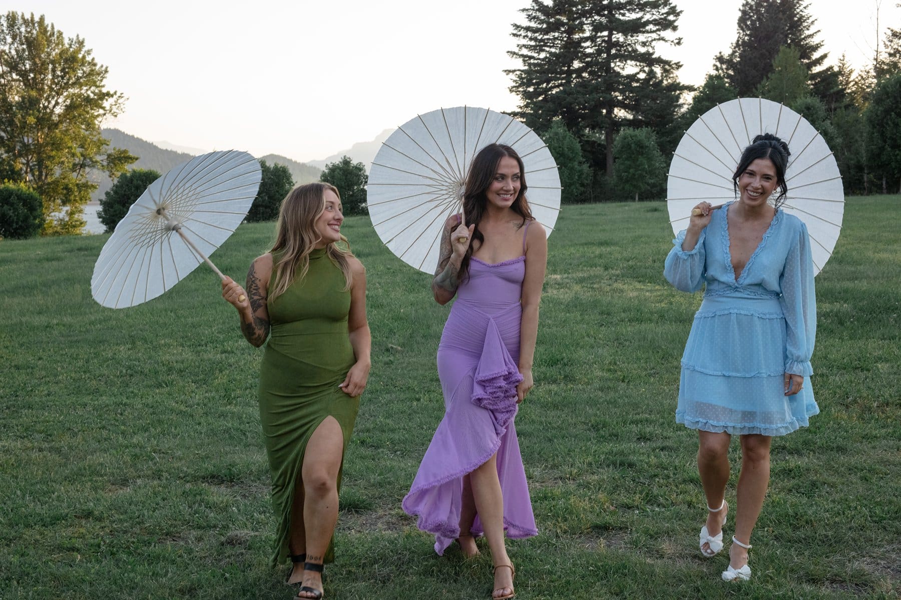Three bridesmaids with parasols laughing and walking across the lawn in colorful dresses at Wind Mountain Ranch.