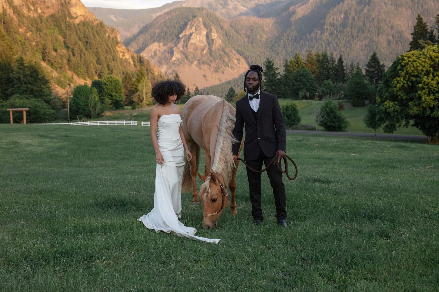 Bride and groom pose with a light tan horse in the open field of Wind Mountain Ranch venue, mountains glowing behind them.