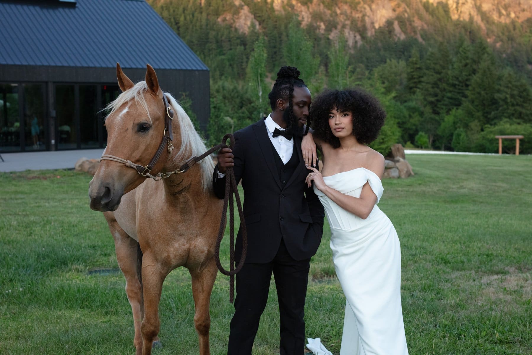 A bride and groom pose with a horse on the lawn at wind mountain ranch venue, with mountains and a modern black reception building in the background.