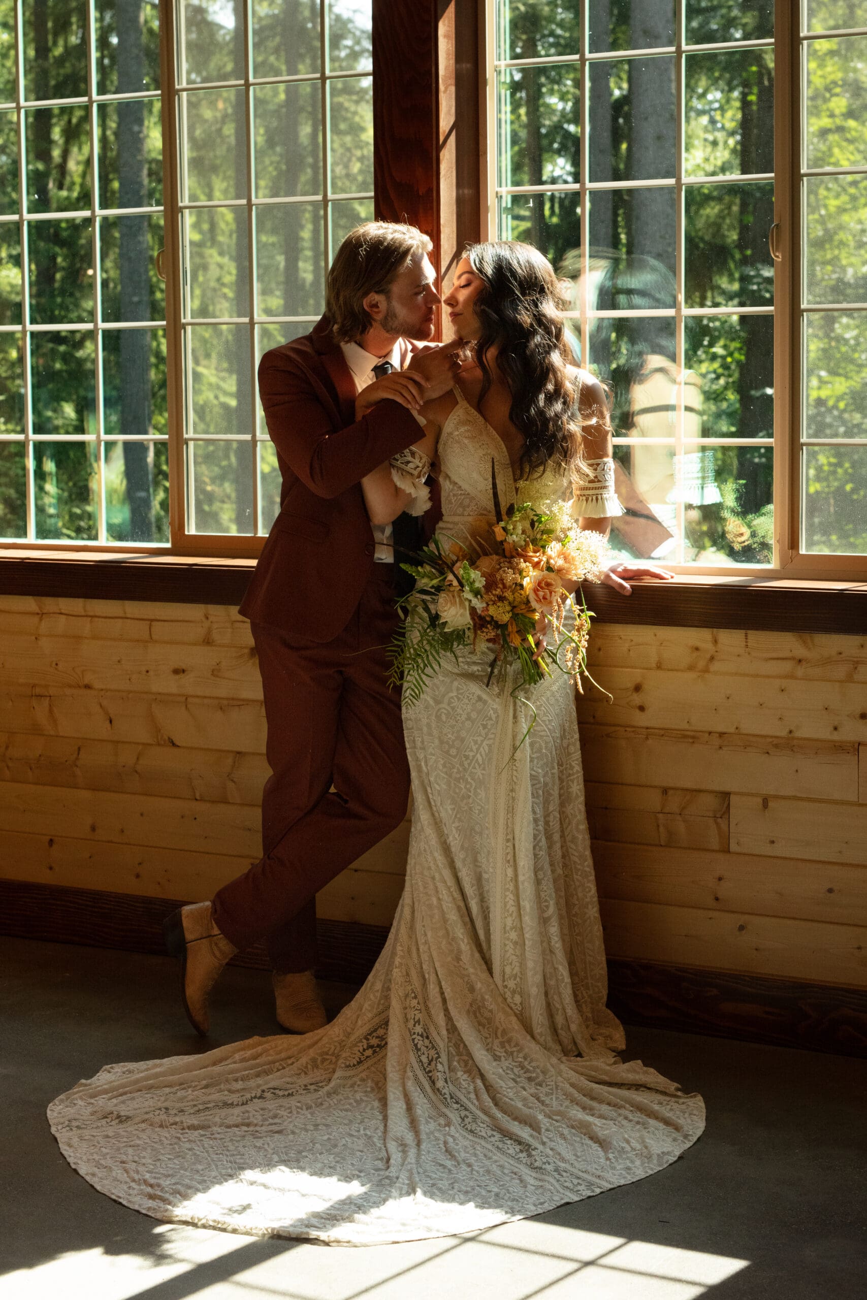 Bride and groom share a quiet moment in sun-drenched lodge, bouquet resting on window ledge – cozy indoor ideas for weddings in the forest.