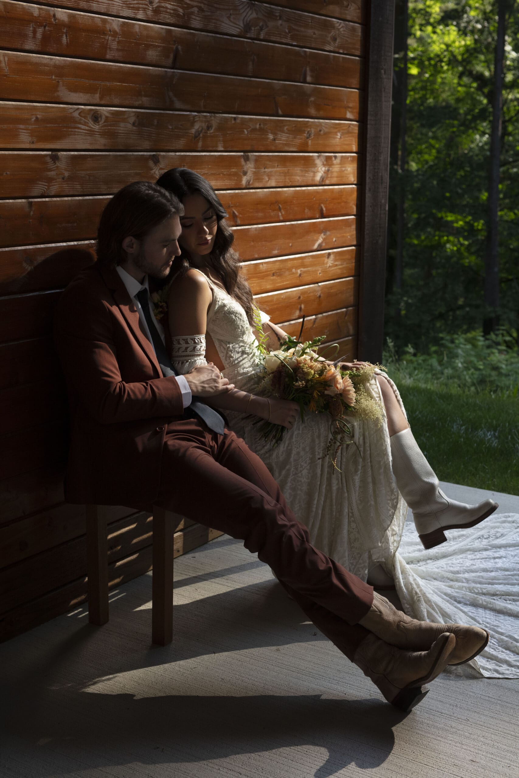 Bride in cowboy boots sits with her groom in evening light beside a rustic cabin – western-inspired ideas for weddings.
