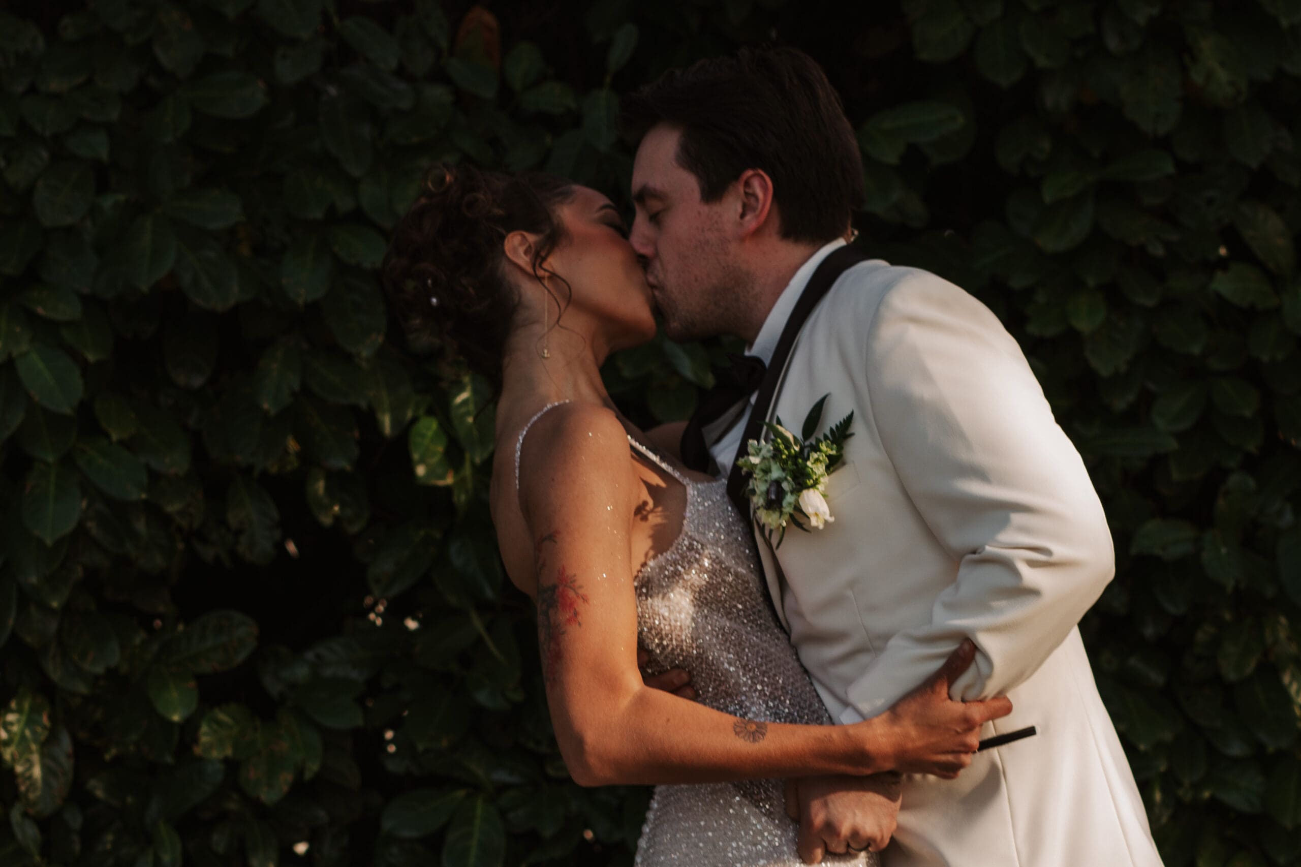 Bride and groom share a kiss in front of lush greenery, showing how bold fashion and candid moments make great ideas for weddings.